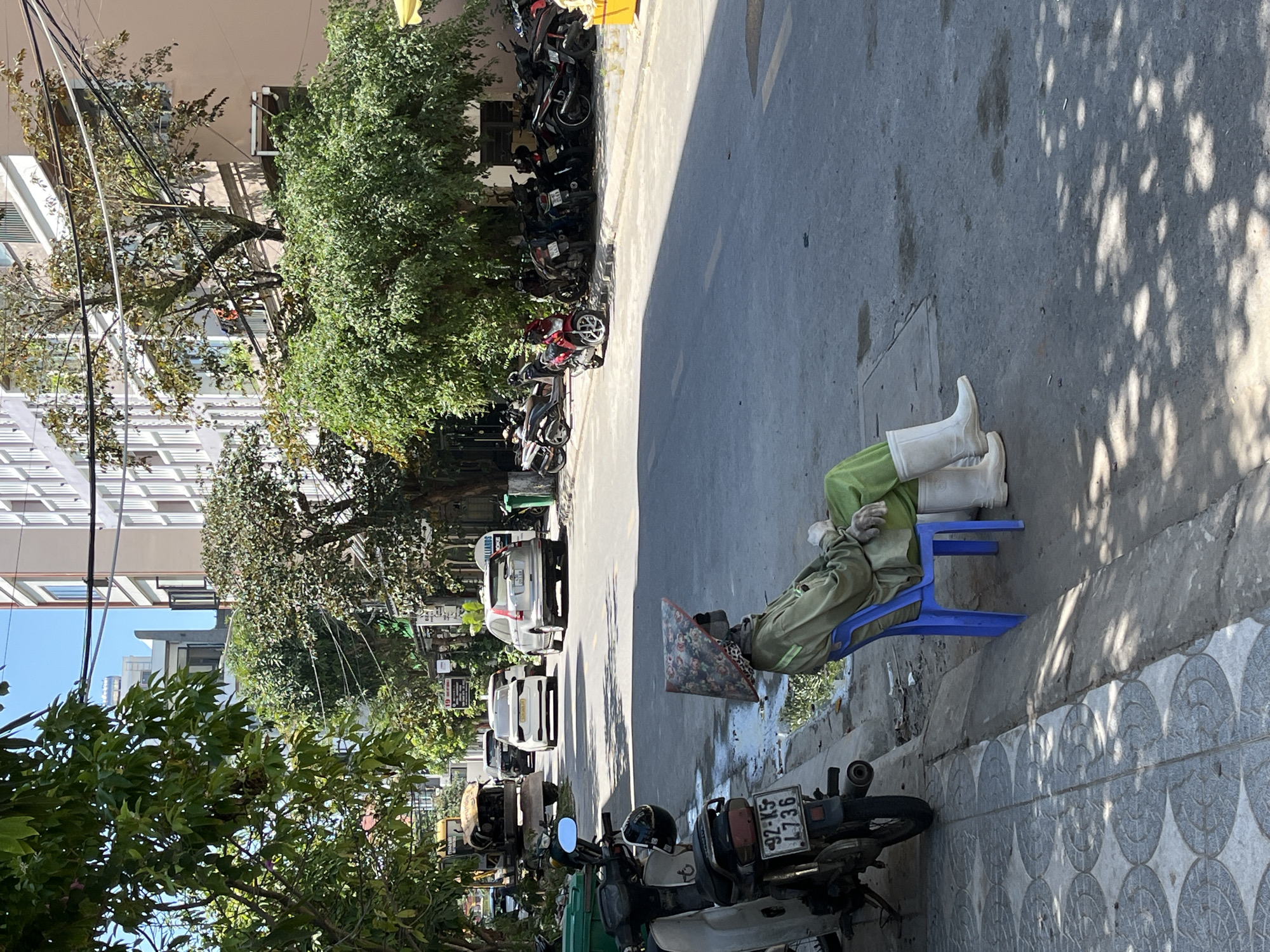A medium shot depicts a person seated on a small blue plastic chair on the side of a street. The person is wearing long-sleeved green attire, white gloves, and white boots. A conical hat is placed near them on the ground. The scene unfolds in an urban setting, with parked motorcycles, cars, and lush green trees lining the street. Tall buildings are visible in the background. The license plate of a motorbike reads "42-K5 4736," which is a typical license plate in the province of Kon Tum, Vietnam. Since Kon Tum has the number 82, most likely it's in the city of Đà Nẵng, Vietnam.