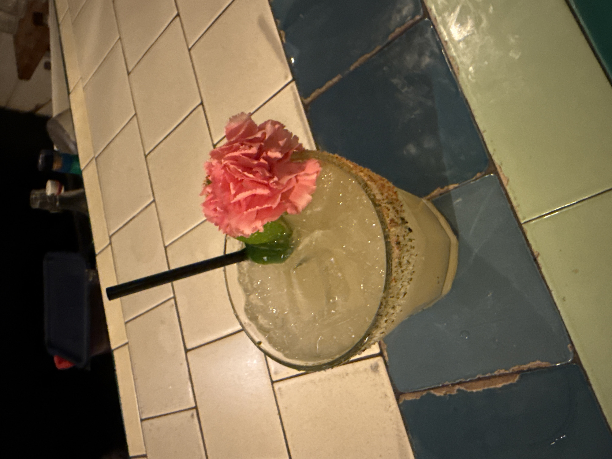 The image captures a cocktail, likely a margarita, placed on a tiled surface composed of white and bluish-green tiles. The cocktail is in a glass with a rim coated in a granular substance, possibly salt or sugar. It is adorned with a pink flower and a green lime wedge, and a black straw extends from the drink. The tiles appear to be somewhat aged, with visible wear and tear. In the background, slightly out of focus, are what seem to be bottles and a dark object. The scene is set in Los Angeles, United States of America.