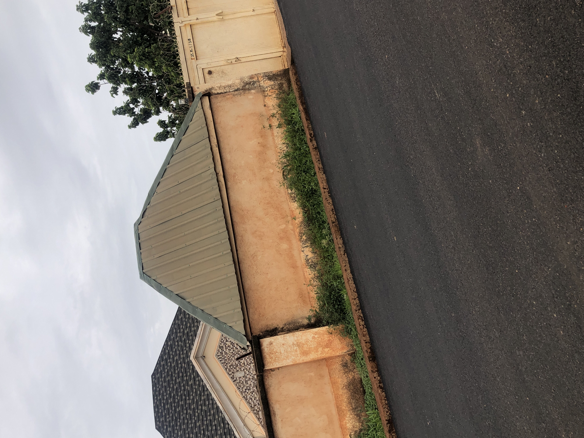 The image displays a street scene with a focus on a residential property. In the foreground, a newly paved asphalt road slopes upwards. Adjacent to the road is a raised flower bed or border with some overgrown green vegetation. Behind this, a peach-colored wall with dark staining at the base extends across the frame. To the left, a portion of a house roof with dark shingles is visible, along with a decorative tiled section of the building. A green corrugated metal roof forms a prominent triangular shape over the wall. To the right, a large cream-colored metal gate with a barbed wire extension on top is set into the wall. The gate has a visible inscription above it. In the background, lush green trees are present. The sky is overcast with gray clouds.