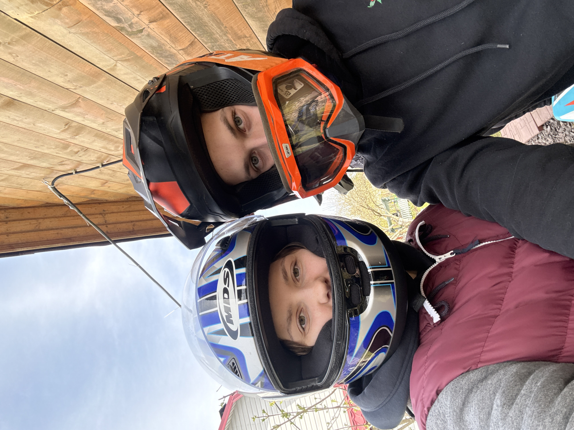 Two individuals wearing motorcycle helmets are positioned close together. The person on the top wears a black helmet with orange accents and orange goggles. The person below wears a silver and blue motorcycle helmet. They are outdoors, with a wooden structure above the person with the orange helmet and a glimpse of the sky and trees in the background. The person on the bottom is wearing a burgundy vest over a grey sweatshirt. The specific location is not identifiable as Ленинградская, Russia based on the given image.
