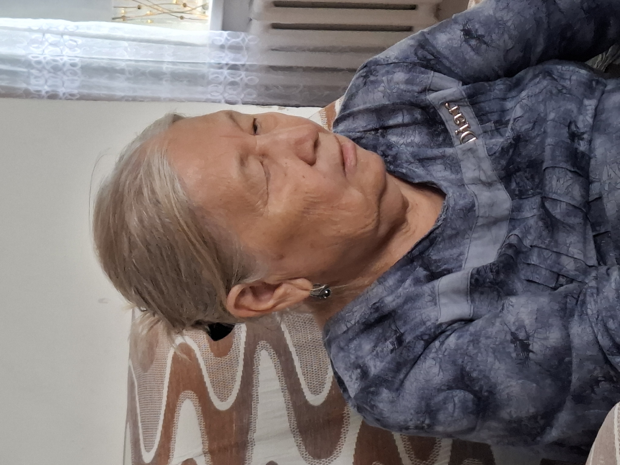 The image shows an elderly East Asian woman sitting in a chair indoors. She has grey hair pulled back and is wearing a blue, patterned top with a decorative strip across the chest that has the word "Diar" on it. She is looking down and to the right. In the background, there is a window with white curtains and a radiator. A chair with a brown and white patterned upholstery is visible on the left. The setting appears to be a residential interior. This image does not contain information that would allow for the determination of its location within the city of Almaty, Kazakhstan.