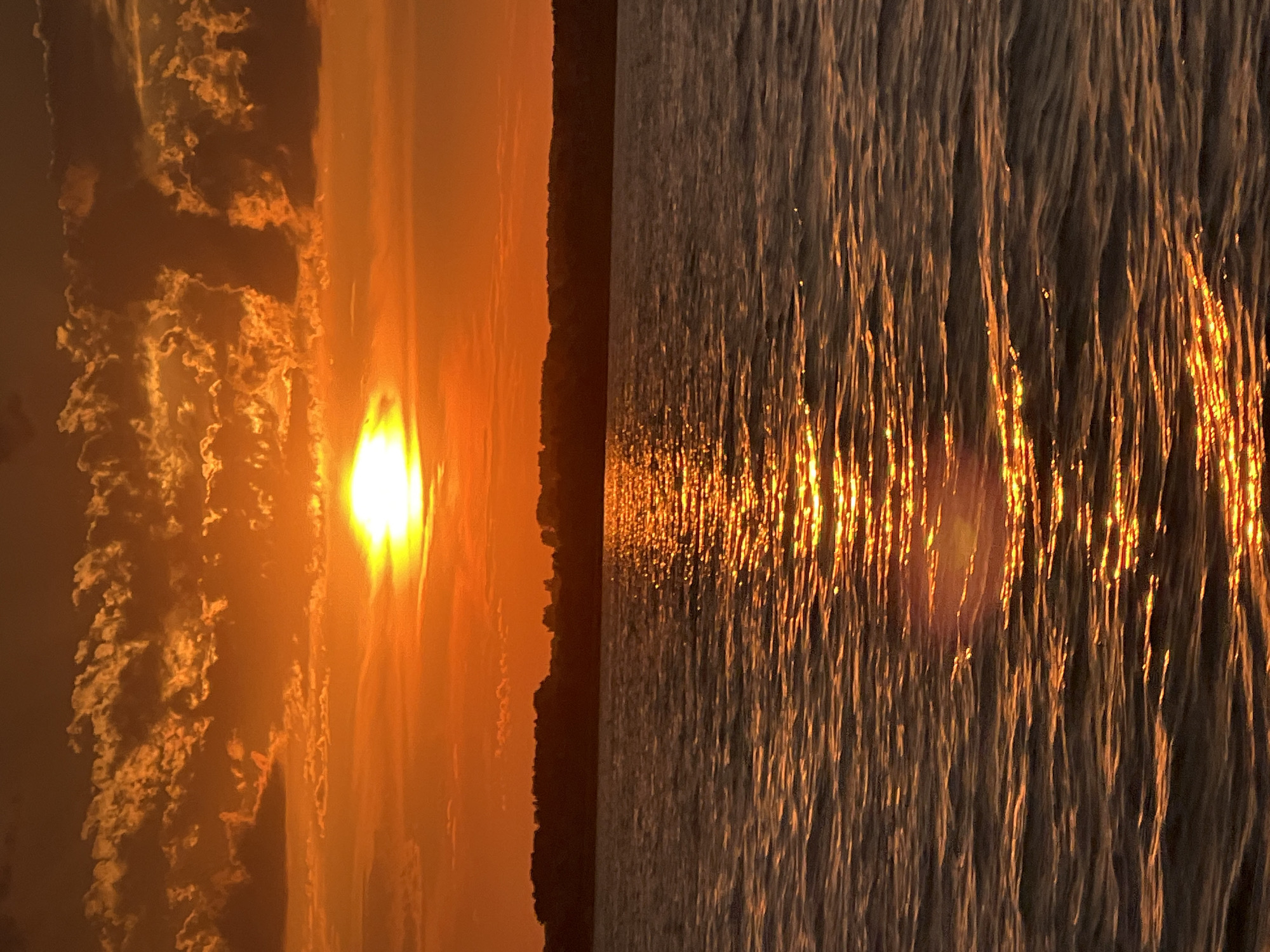 This is a photograph of a sunset over a body of water, taken from a boat on the Ob River in Novopichugovo, Russia. The sun is low on the horizon, casting a warm, orange glow across the sky and reflecting on the rippling water. The sky is filled with dramatic clouds, illuminated by the setting sun, and a silhouetted treeline marks the far bank of the river. The water is dark, with bright streaks of sunlight creating a shimmering effect. There are no people or specific objects visible in the image, only the natural landscape.