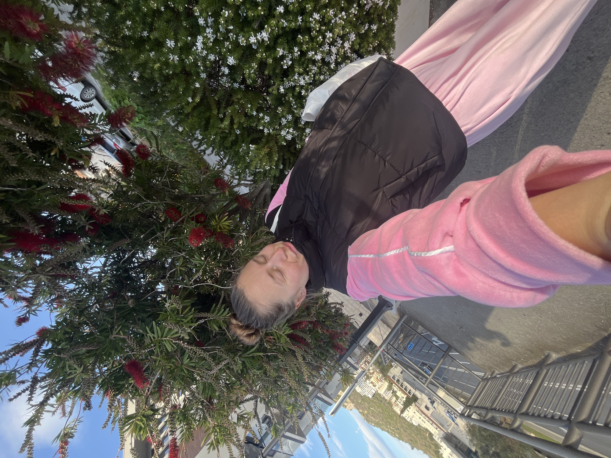 A person wearing a pink tracksuit and a black vest is lying down next to a tree with red, bottlebrush-like flowers. The person's eyes are closed and their hair is in a bun. The location is Boreti, Montenegro, evident from the buildings and landscape in the background. A metal railing and road are visible behind the person.