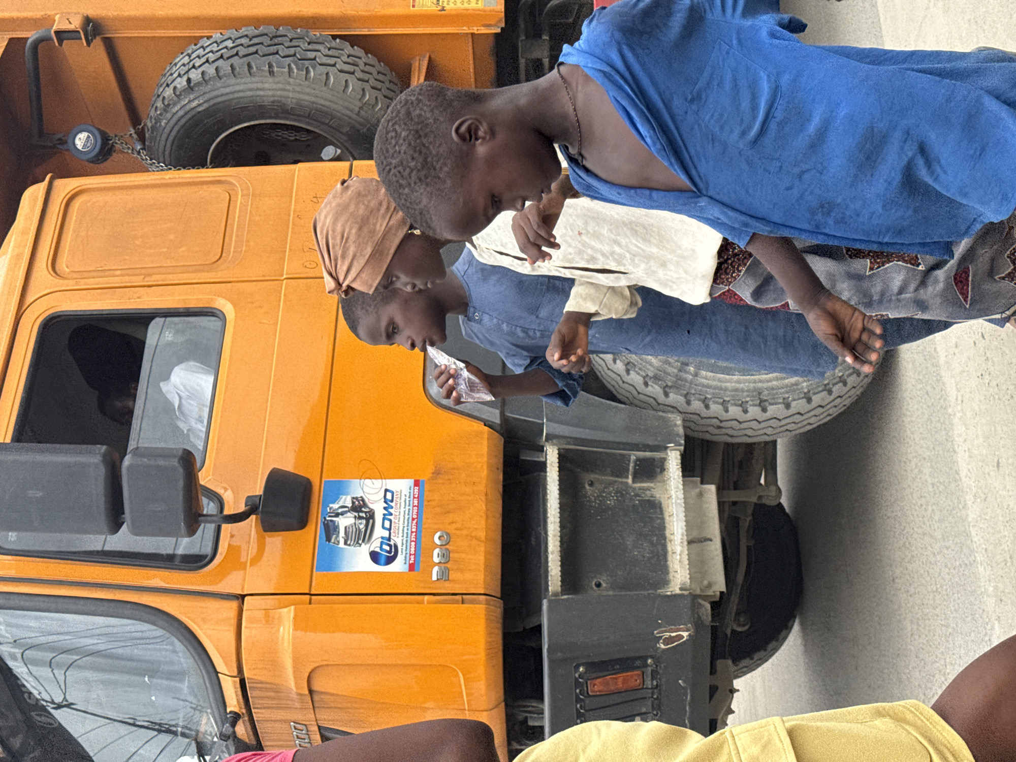 In Ado, Nigeria, a bright orange dump truck is parked on a dusty, light-colored road. Three young children stand to the left of the truck. The child in the foreground, wearing a bright blue shirt, looks down and to the right, with their right arm extended. Behind them, a child in a white shirt and patterned pants looks down and to the left, with their left hand holding a plastic bottle of water up to their mouth. The third child, furthest back and to the left, wears a blue garment and is also looking to the left. A spare tire is visible mounted on the side of the truck. A logo for "OLOWO GROUP OF COMPANY" with a picture of a truck is affixed to the side of the truck, along with the number "380". The truck's cab has a visible driver inside.