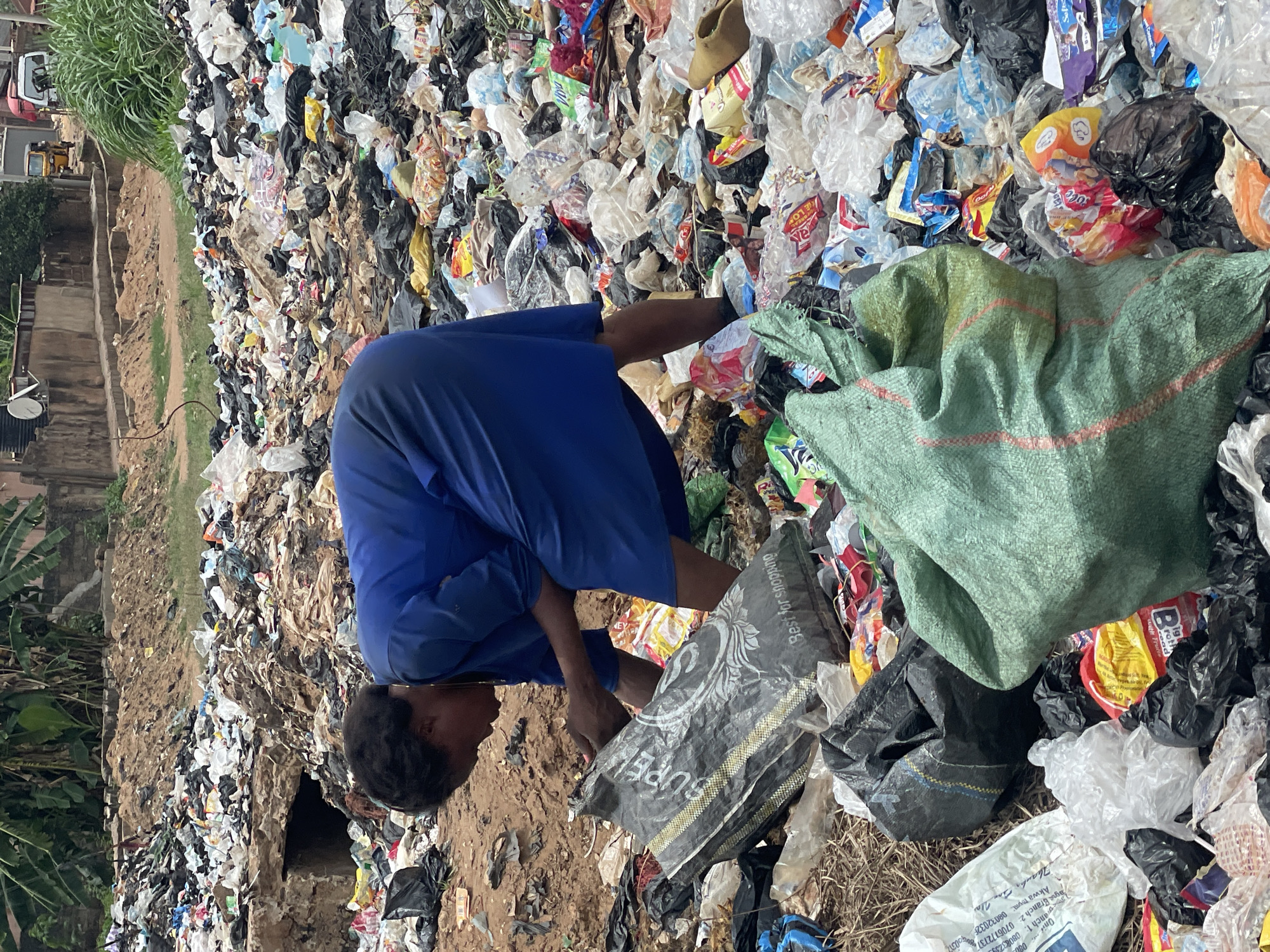 The image depicts a large, unsanitary landfill with a man scavenging through the trash. The man is wearing a blue shirt and is bent over, sorting through the waste. There are numerous plastic bags, food wrappers, and other refuse scattered around him. The scene is in Onitsha, Nigeria, as evidenced by the writing on one of the discarded plastic bags. There is a hill of trash to the left of the man and a green burlap sack near his right.