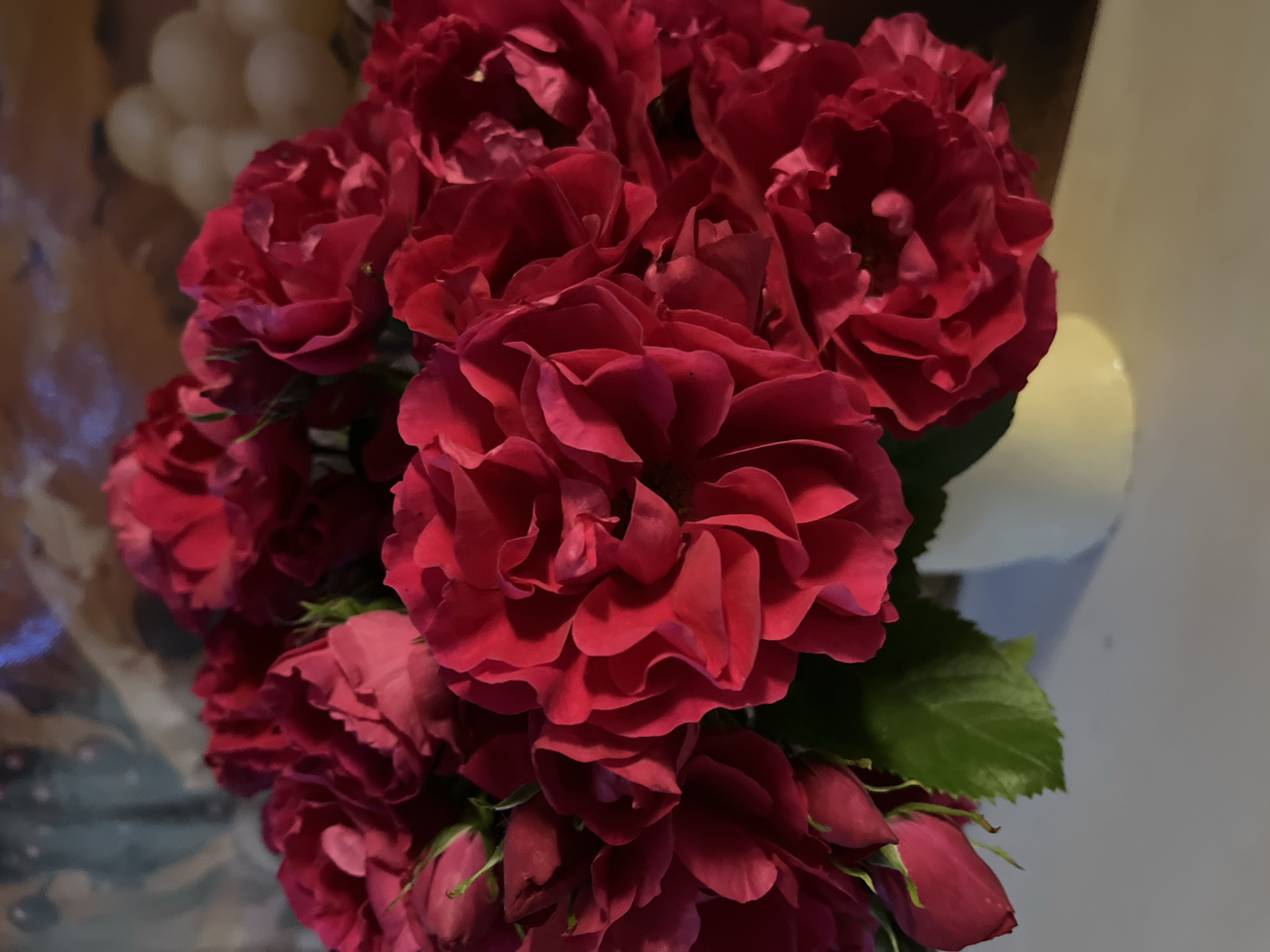A close-up, overhead shot captures a bouquet of vibrant red roses in a white vase. The roses are densely packed, with their velvety petals unfurling in intricate patterns. Some buds are visible amongst the blooms, hinting at their potential. The background is softly blurred, revealing hints of light-colored, rounded objects, possibly balloons, and a darker, textured surface. The scene appears to be indoors, with the bouquet resting on a light-colored surface. The image has a warm, intimate feel, with the lighting casting subtle shadows and highlighting the rich color of the roses. There is no indication of the city of Kalynivka, Ukraine, or any people present in this image.