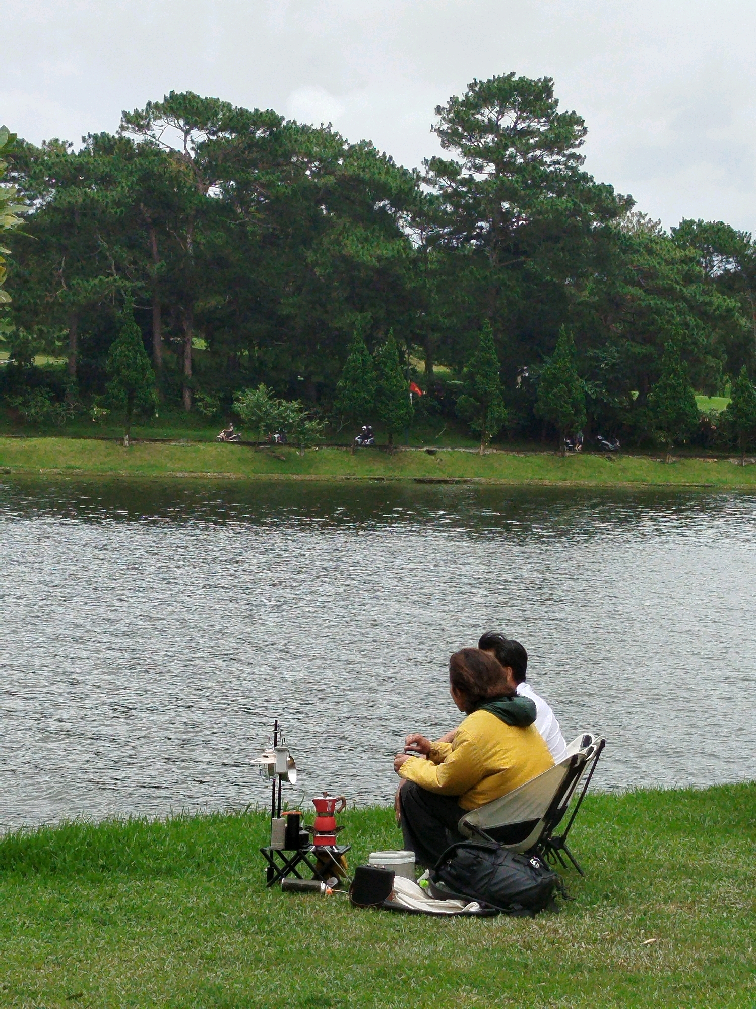 In Da Lat, Vietnam, two individuals are seated on foldable chairs on a grassy bank beside a calm body of water. The person in the foreground, wearing a yellow jacket and black pants, faces away from the camera. To their right, a second individual in a white shirt is also seated. In front of them, a small portable stove with a red coffee pot and other cooking equipment is set up on a small table. A black backpack rests on the ground near the chairs. The background features a dense line of pine trees that extend across the horizon, bordering the water. The sky is overcast. Further in the background, across the water and beyond the trees, more trees and some structures are visible. In the distance, along the treeline, there are indications of other people, possibly on motorbikes or scooters.