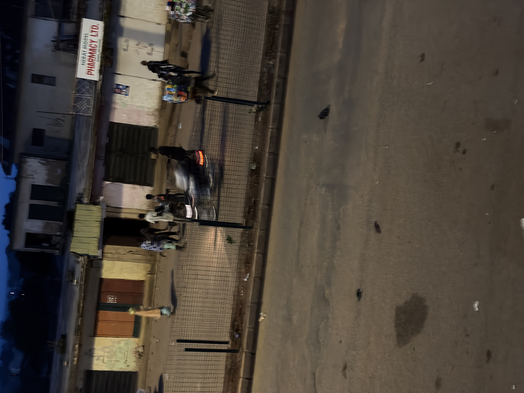 This image captures a street scene in Bukuru, Nigeria, likely during the evening or early morning given the low light. The perspective is from above, looking down at an angle.
On the left side of the frame is a row of buildings with storefronts. One of these storefronts has a prominent sign reading "NERAT MODEL PHARMACY LTD.". The buildings appear somewhat aged, with some showing signs of wear. Several people are visible on the sidewalk in front of the buildings, some walking, others gathered in small groups. One person is pushing a cart loaded with what appear to be bottles.
The street itself occupies the right side of the image. It is paved and appears to be a two-way road. There is a distinct curb separating the sidewalk from the street. A dark-colored vehicle, possibly a car or motorcycle, is moving along the street, with its brake lights illuminated. Shadows are cast, indicating a light source from above and to the left.
The overall atmosphere seems to be that of a typical urban street with pedestrian activity and some vehicular traffic. The lighting suggests a time of day when artificial lights are on or when natural light is dim. There are no strong indicators of weather conditions, but the pavement appears dry. The scene depicts everyday life on a street in Bukuru.