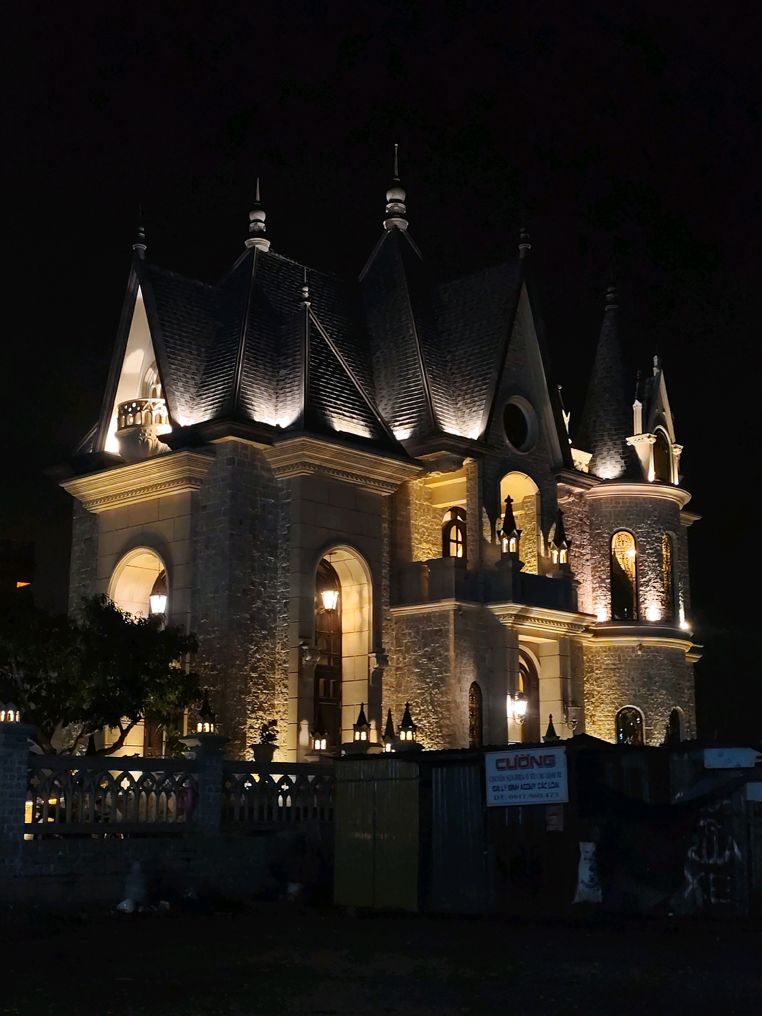 The image shows a large, ornate building with a gothic architectural style, illuminated at night. The building features multiple spires, arched windows, and stone walls. It appears to be a mansion or a similar structure. In front of the building is a decorative fence and some trees. In the foreground, there are some corrugated metal structures and debris on the ground, suggesting an area under construction or an informal settlement. A sign with Vietnamese text is visible on one of the metal structures. The scene is set at night, with the building being the primary light source.
This building is identified as "Dinh 1" or "Dinh Bảo Đại" in Da Lat, Vietnam. It was the former residence of Emperor Bảo Đại, the last emperor of Vietnam. The architectural style is a blend of French colonial and local influences, with Gothic elements being prominent in the design. The current state of the area in front of the mansion suggests ongoing renovation or redevelopment.