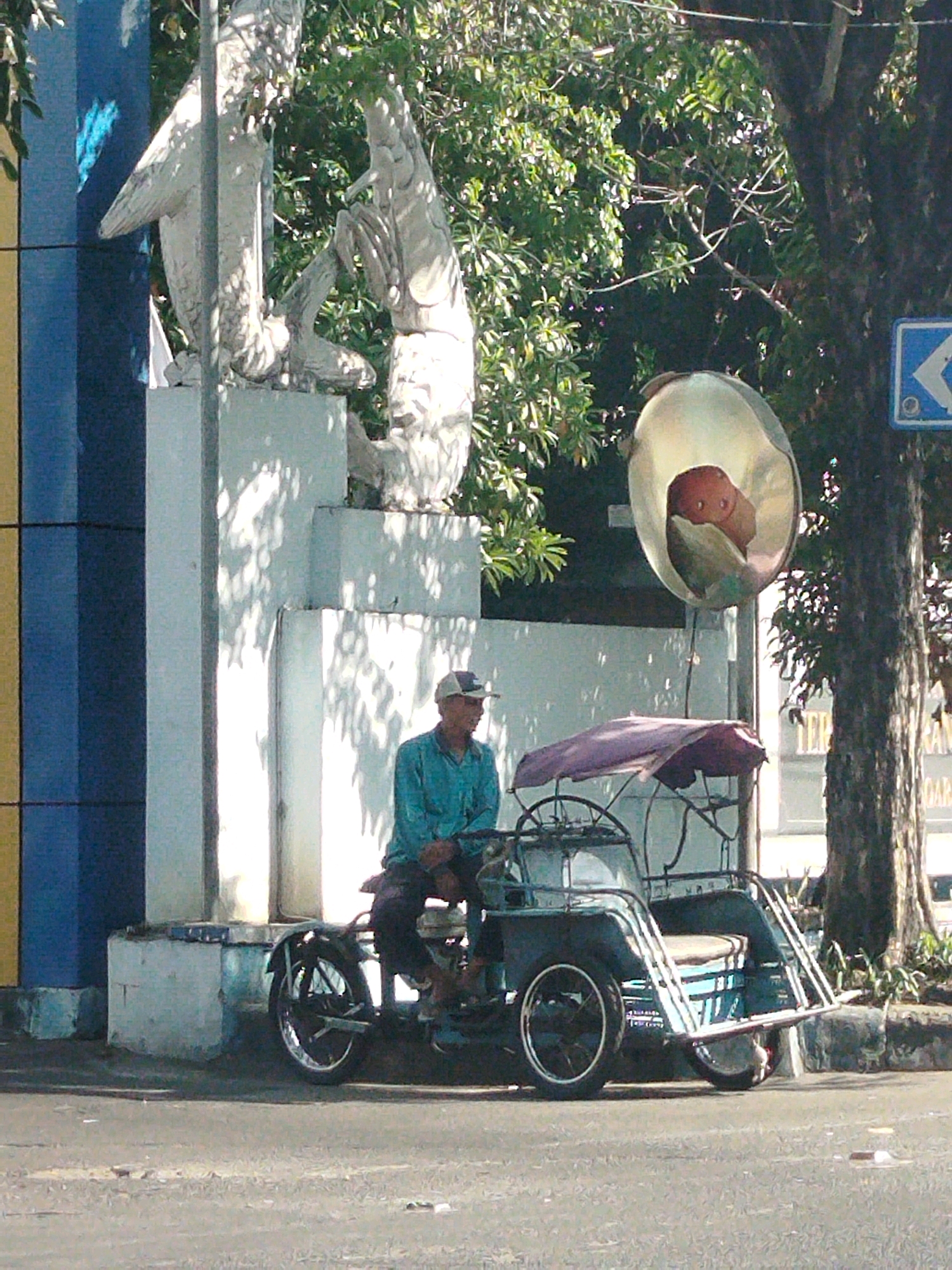 The image depicts a man in a turquoise shirt and a baseball cap sitting in a pedicab, which is a three-wheeled vehicle powered by a person pedaling. The pedicab has a purple canopy and is parked on a street. Behind the pedicab, a large tree with dense foliage is visible, and to the left, there are large, white concrete structures, possibly part of a monument or building. A sign with a blue background and a white arrow pointing left is also present. The scene appears to be outdoors during the daytime, with sunlight casting shadows on the ground and structures. The location is identified as Larangan, Indonesia.