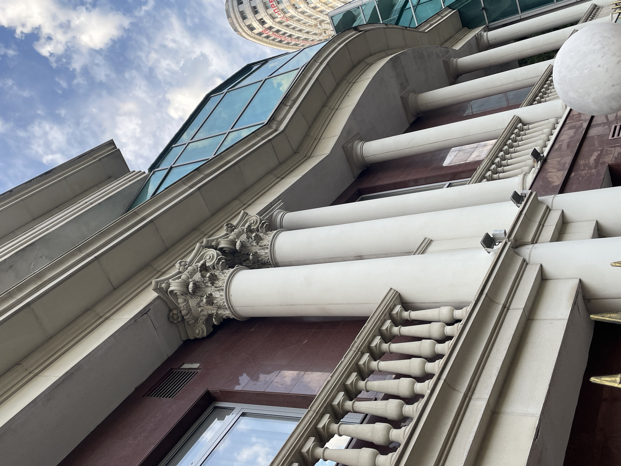 This media file is an upward-facing, wide-angle shot of an ornate building with classical architectural elements. The subjects are the building's facade and the sky above. The building features a dark red marble exterior, large white columns, and a decorative stone balustrade. A modern glass-paneled section of the building curves upwards, reflecting the sky and clouds. In the distance, another curved building with many windows is visible against the sky.
The scene is set outdoors, likely in an urban environment given the presence of large buildings. The time of day appears to be daytime, with a bright sky filled with scattered white clouds, suggesting fair weather. There are no people visible in the media file.
Notable details include the detailed stone carvings on the columns and the repetitive pattern of the balustrade. The strong upward angle creates a sense of grandeur and height. The contrast between the traditional architectural style of the lower part of the building and the modern glass structure highlights different design periods. No text is clearly visible in the image. The context provided indicates the location is Sochi, Russia.