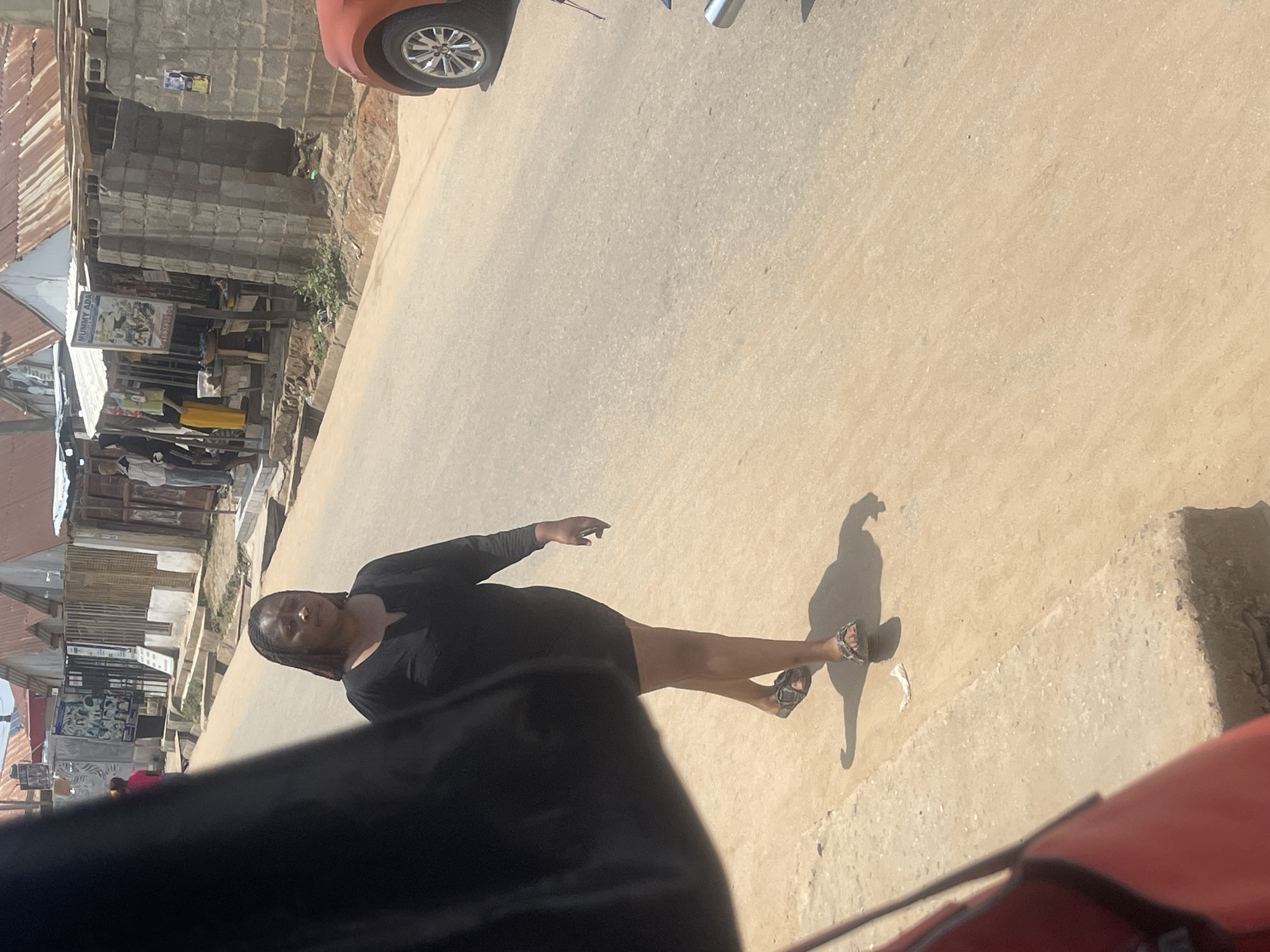 A woman in a black dress walks on the right side of a dusty road in Abuja, Nigeria. She is wearing sandals. Behind her, buildings with corrugated iron roofs line the street. An orange car is parked on the right side of the road. A black bag is in the foreground on the left side of the image.