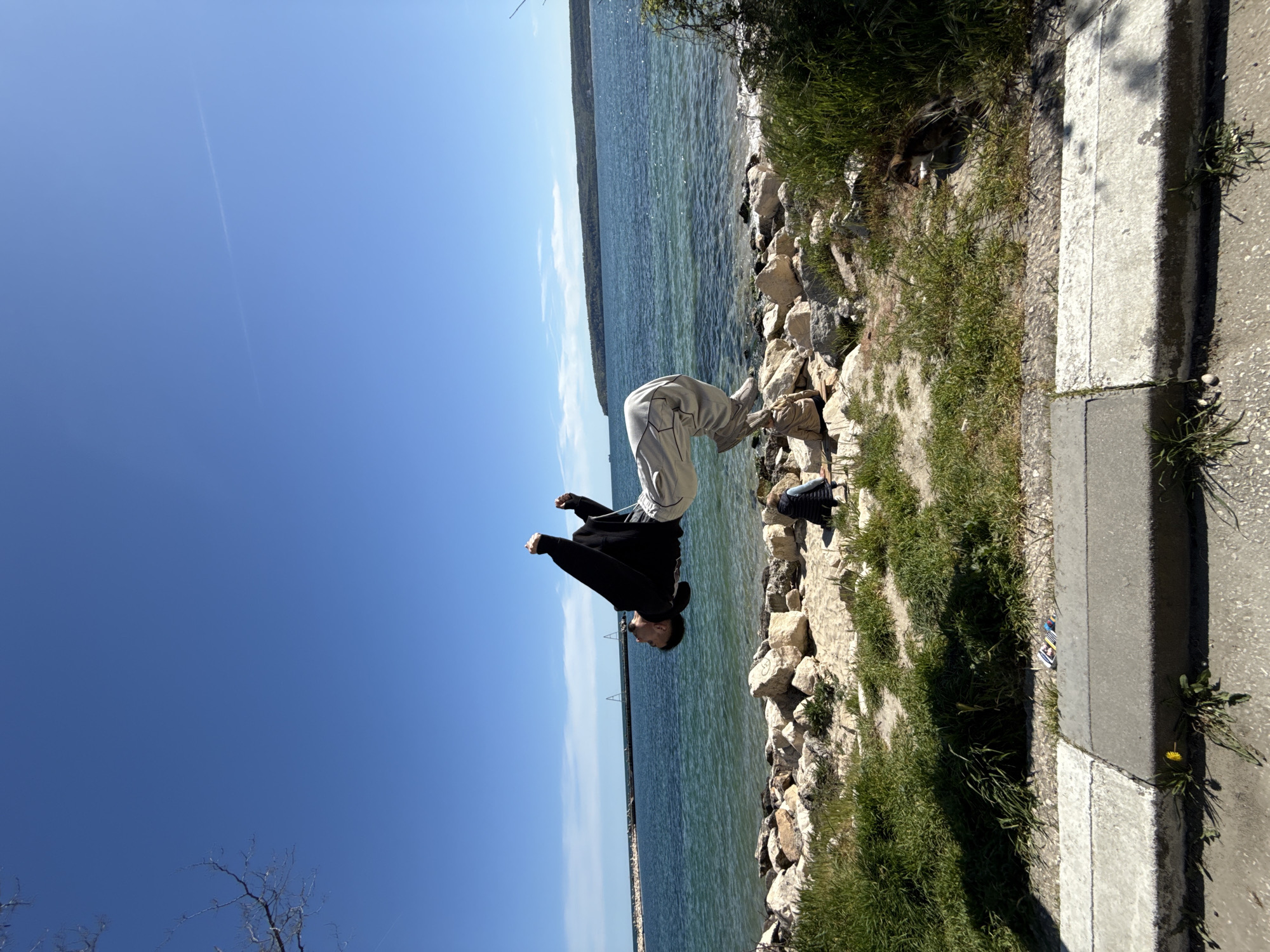 This image captures an athletic individual performing an acrobatic flip on a bright, sunny day by the sea in Varna, Bulgaria. The person, dressed in a black long-sleeved top and light-colored pants, is suspended mid-air in an inverted position, with their head pointing towards the water and their feet upwards, hands clenched.
The setting features a clear, turquoise-blue body of water, likely the Black Sea, on the left side, with a distant, tree-covered landmass visible across the horizon. The sky above is a brilliant, cloudless blue, with a faint contrail marking it. The foreground comprises a rocky shore adjacent to the water, which transitions into patches of green grass. On the far right, a concrete curb separates the natural landscape from a paved surface, possibly a sidewalk or road. Sunlight casts distinct shadows on the ground, indicating midday or early afternoon. A small yellow flower can be seen growing near the curb in the grass. No visible text is present.