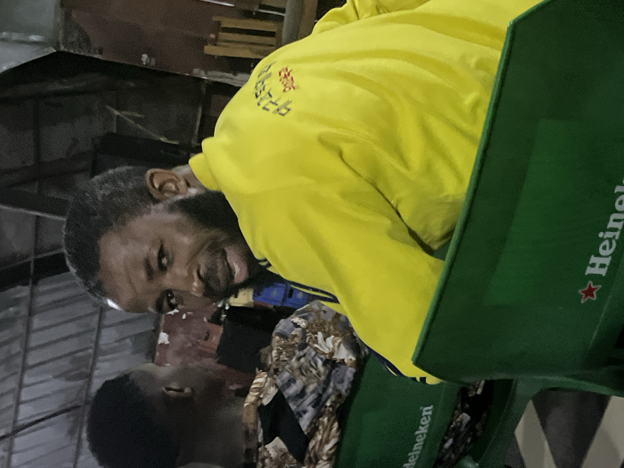 In the city of Jos, Nigeria, two men are seated in a dimly lit indoor setting, possibly a bar or restaurant. The man in the foreground, facing away from the camera, wears a bright yellow jacket with dark blue stripes on the sleeves and a red and blue embroidered logo on the back. He has dark skin, short, dark hair, and a beard. The man to his left, facing the camera, is partially obscured but appears to have dark skin and short, dark hair. He is looking directly at the camera with a slight smile. They are seated on green plastic chairs with the Heineken logo visible on one of them. The background is dark and shows some wooden structures and a corrugated metal roof. The overall atmosphere is casual and relaxed.