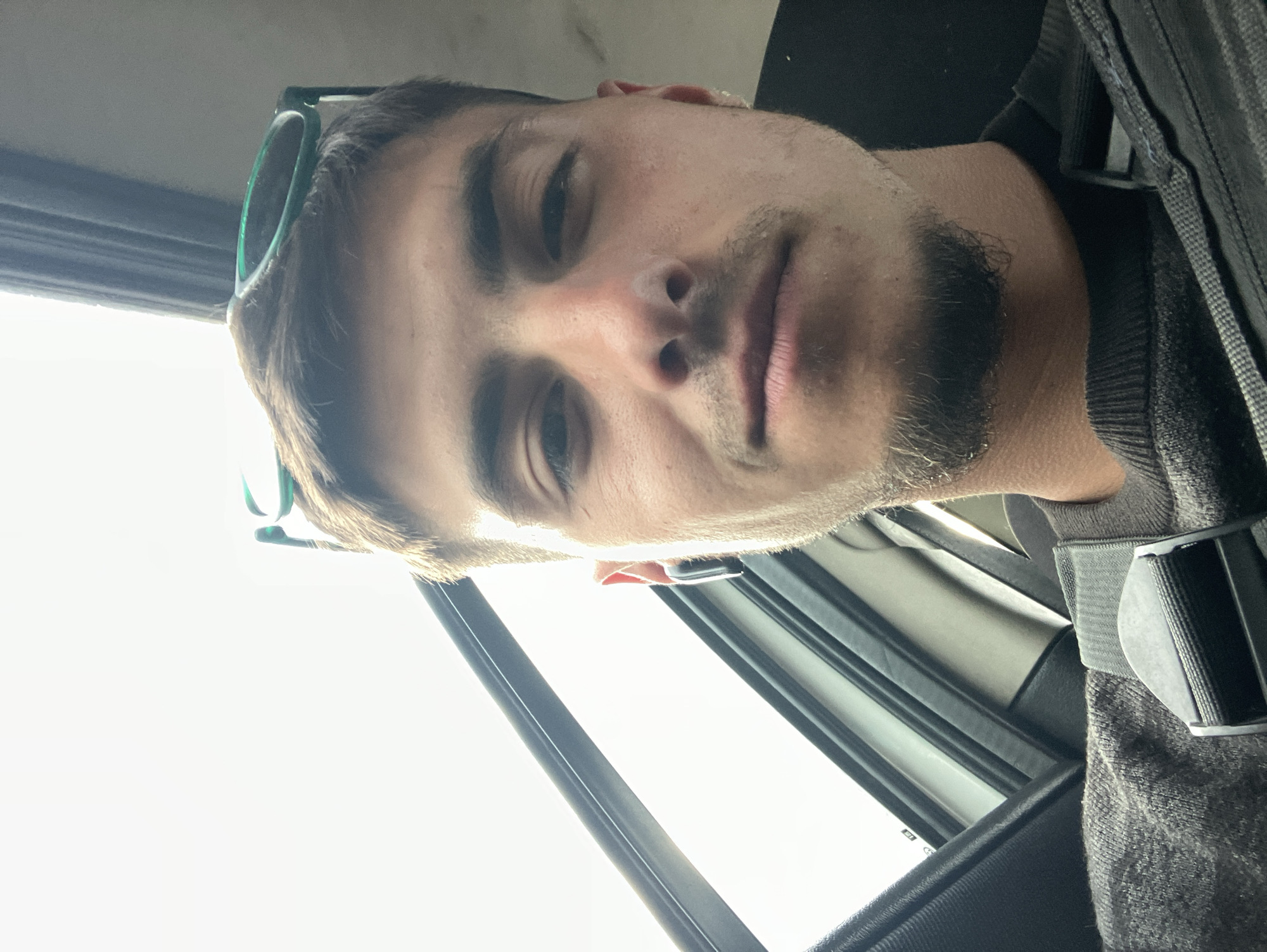 A close-up selfie of a young man with a light complexion, brown hair, and a trimmed beard. He is wearing a gray sweater and a seatbelt. Green sunglasses are perched on top of his head. The background shows the interior of a car with a window. The location is likely Yoakim Gruevo, Bulgaria.