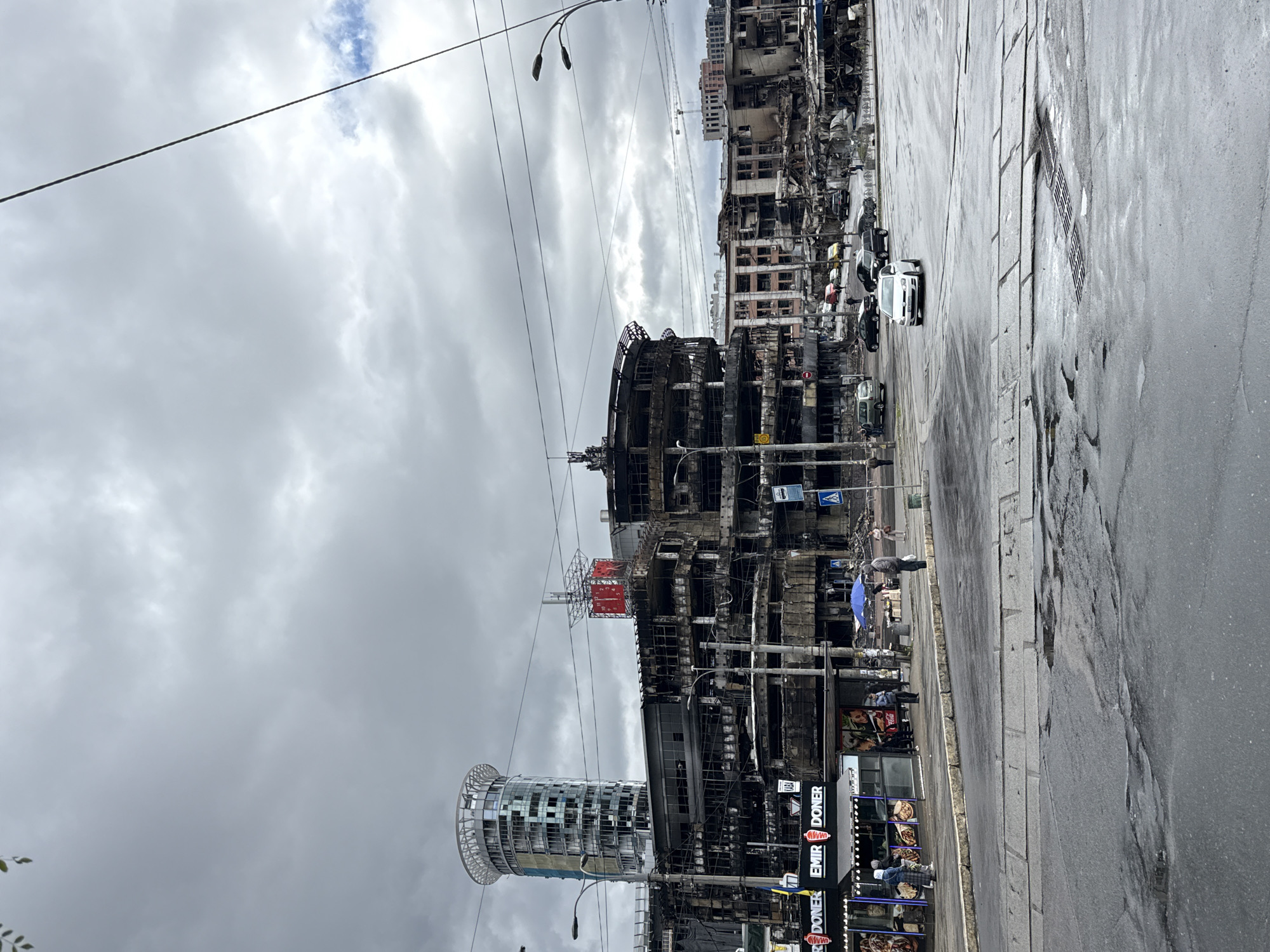 The image depicts a street scene in Kyiv, Ukraine, showing buildings that have been heavily damaged, apparently by fire. The skeletal remains of multi-story structures are visible, with their interiors exposed. A modern glass-facade building stands adjacent to the destroyed buildings, seemingly intact. On the street level, there is a food stand, with a sign that says "Emir Doner". Several people are walking or standing along the sidewalk. Cars are parked along the street. The sky is overcast, and the street surface appears wet.