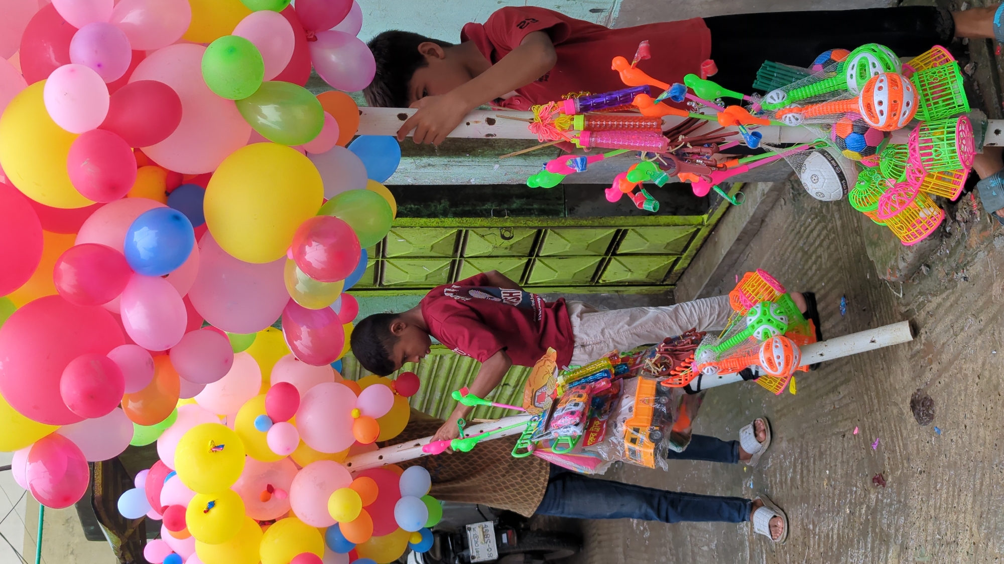 Two young boys are standing on a wet, textured city sidewalk in front of a lime green, metal barred door in Dhaka, Bangladesh. The boy on the left, wearing a red t-shirt and white pants, is holding a pole with balloons and toys. The boy on the right, also wearing a red t-shirt, is holding a pole with colorful toys and balloons. The sidewalk is wet and appears to have some puddles. There are various colorful toys, including toy trucks, bubble blowers, and plastic cages, displayed on poles. Numerous colorful balloons are also displayed overhead. A motorcycle is parked behind the boy on the left. The scene appears to be a street vendor selling toys and balloons.
