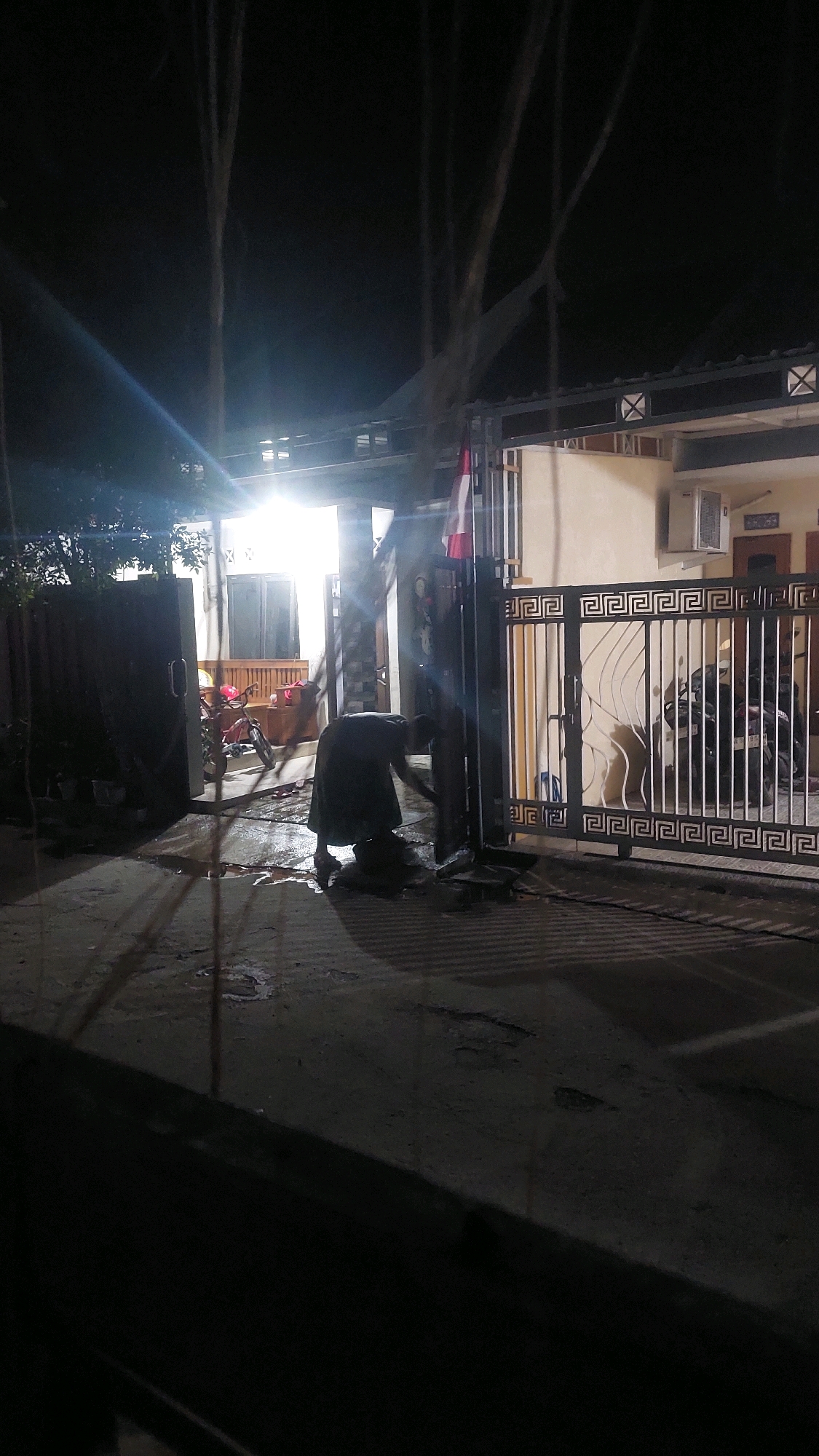 A person wearing dark clothing is bent over and sweeping debris on a wet, paved surface in front of a metal gate. The gate is ornate with a Greek key pattern and has a flag pole with a flag. Behind the gate is a house with an air conditioning unit mounted on the exterior wall. A motorcycle is visible inside the gate. To the left of the gate is a dark, closed gate. A red motorcycle is parked in front of the house, to the left of the main gate. The scene is lit by artificial light, suggesting it is nighttime. The location is identified as Babadan, Indonesia.