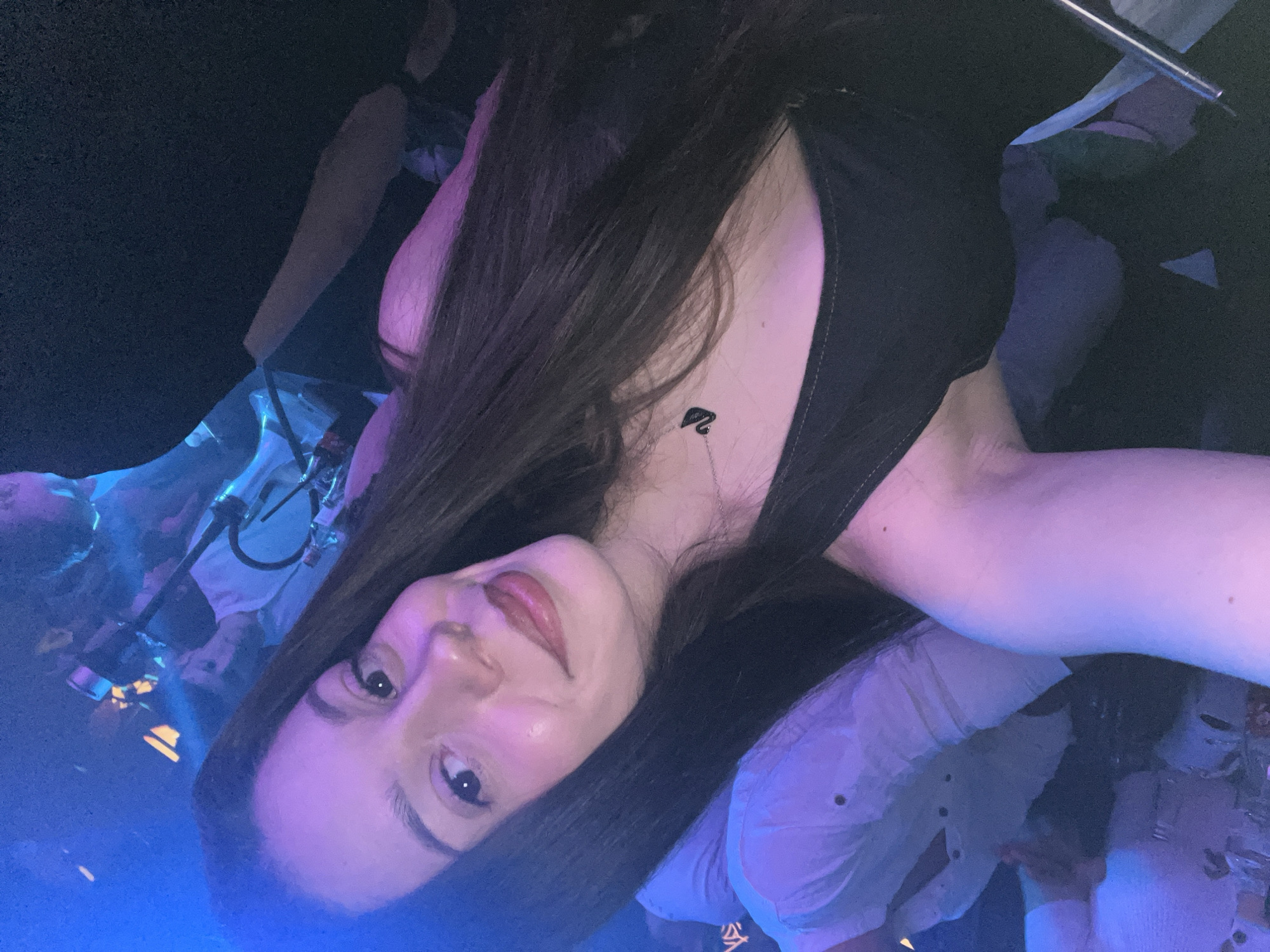 The image depicts a young woman taking a selfie in a club or bar. The lighting is dark, with blue and purple hues dominating the scene. She has long dark hair, fair skin, and is wearing a black top. A small tattoo is visible on her chest. In the background, other people are visible, some wearing white shirts, and what appears to be a hookah or similar device. The location, Sofia, Bulgaria, is not identifiable from the image.