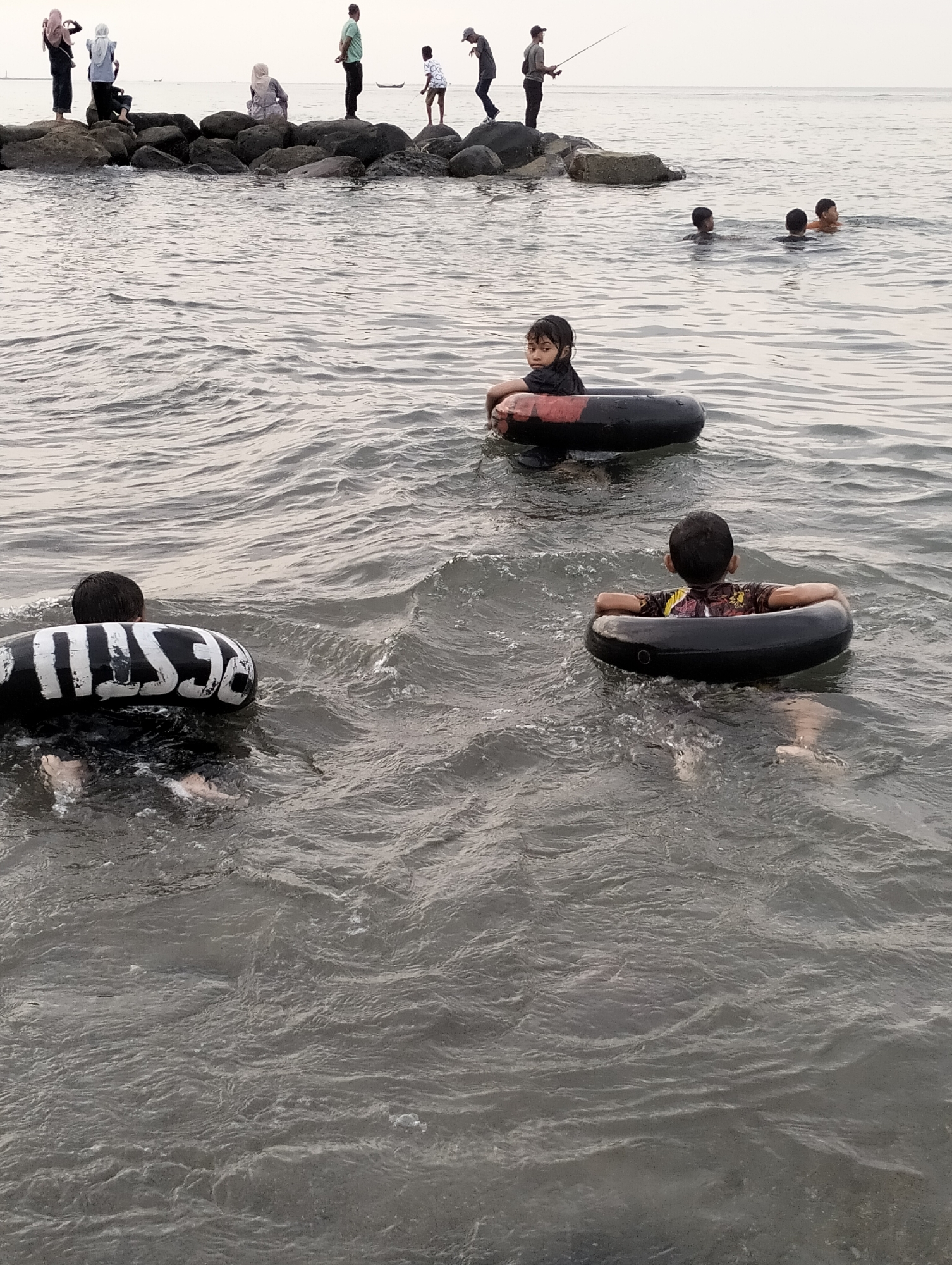The image portrays a coastal scene in Lhokseumawe, Indonesia, featuring individuals engaged in recreational activities. The foreground and midground are dominated by dark, choppy ocean water. Three children are visible in the immediate water: one child in the mid-left faces the camera, wearing a dark shirt and floating in a black inner tube with a red band; another child to the right, facing away, wears a patterned shirt and uses a plain black inner tube; and a third child in the left foreground, partially obscured and facing away, uses a black inner tube with white "BESTIE" lettering. Further back in the water, four additional individuals, likely children, are partially submerged and swimming.
In the background, a rocky breakwater extends horizontally, supporting six individuals. On the left section of the rocks, three individuals, two standing (one with a dark headscarf, one with a light blue top and headscarf) and one seated (with a light headscarf and patterned top), are present. On the right section of the rocks, three men are visible: one standing in a light green shirt, one standing with a fishing rod actively casting into the water, and one standing wearing a cap. The horizon line reveals a light, overcast sky and two small, distant boats on the water. The scene depicts a public waterfront area where people are swimming, fishing, and observing the environment.