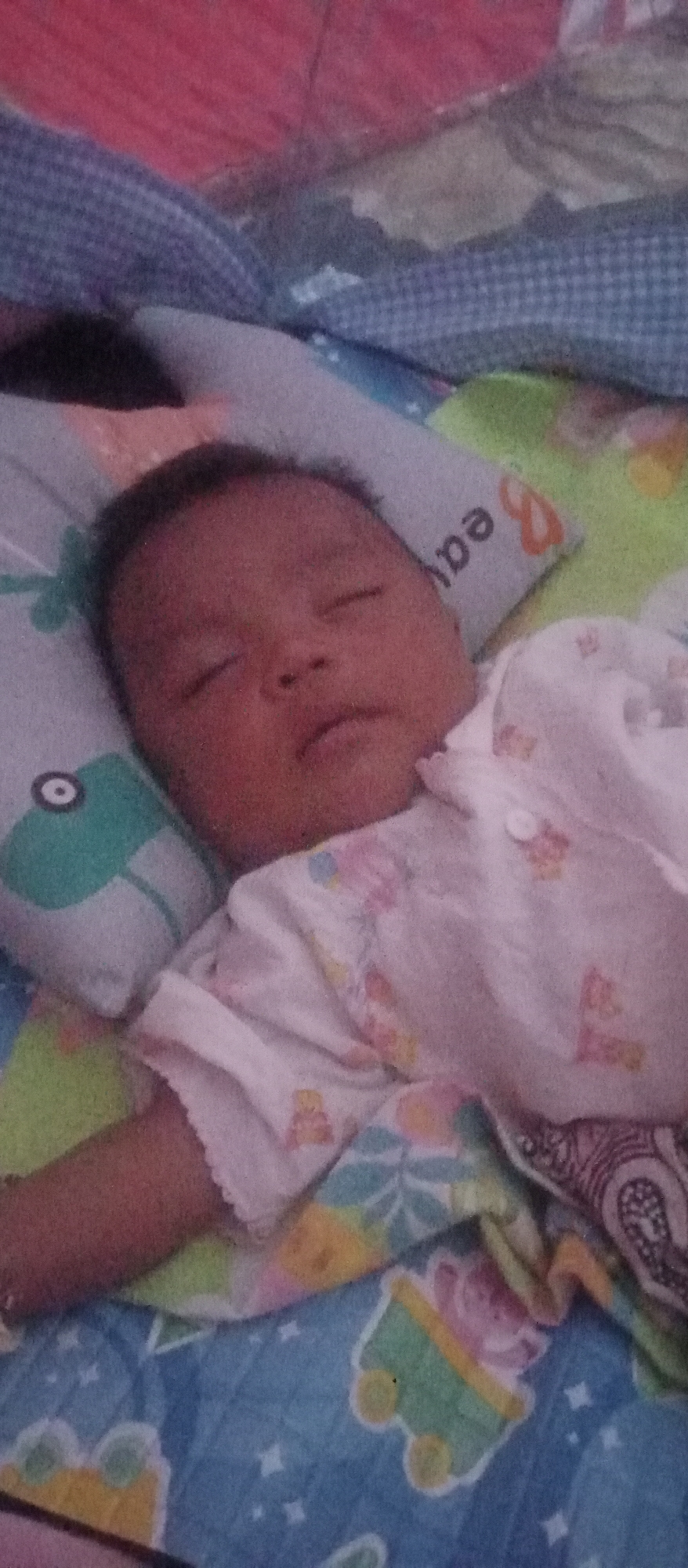 A medium shot captures a baby sleeping on its back on a colorful blanket. The baby is lying on a patterned pillow, facing upwards. The background is a mix of fabric textures, including a pinkish-red material and a blue checkered pattern, suggesting it's on a bed or similar soft surface. The overall scene appears to be indoors, likely in a residential setting in Lubuk Pakam, Indonesia.