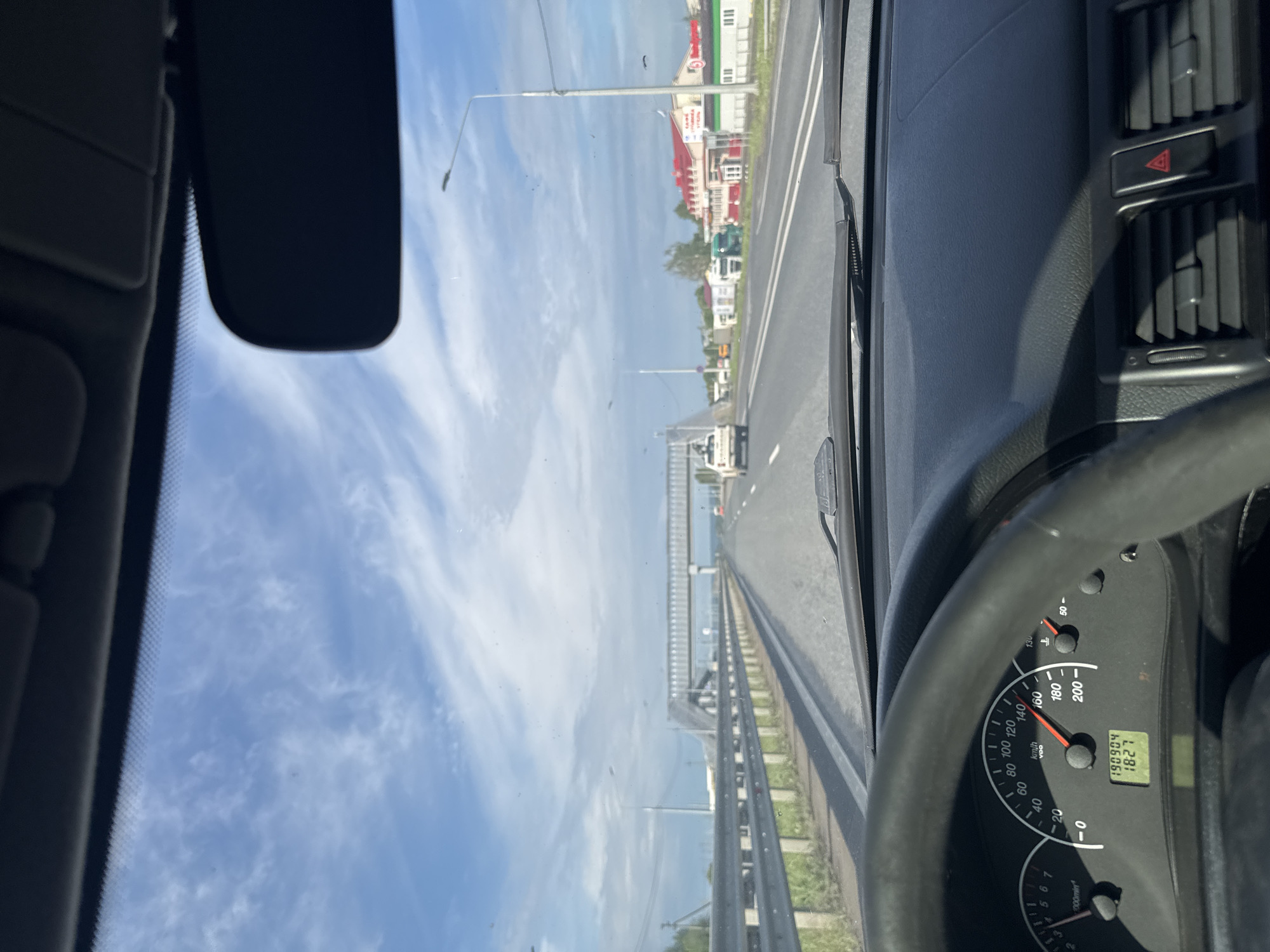 The image captures a driver's perspective from inside a car traveling on a road in Усть-Сыны, Russia. The view through the windshield shows a multi-lane asphalt road stretching into the distance, bordered by a metal guardrail on the right. Beyond the guardrail, what appears to be a body of water or a wide expanse with a distant bridge structure is visible, leading to buildings with colorful rooftops (red and green) and some industrial-looking structures further along the roadside. Overhead, the sky is bright blue with scattered, wispy white clouds. Several streetlights/power poles line the road.
Inside the car, parts of the dashboard and steering wheel are prominent. The black steering wheel occupies the lower right portion. To its left, the instrument cluster is visible, showing a speedometer indicating approximately 80 km/h. An odometer displays "190904" km, and a digital clock shows "18:27". Other text on the speedometer includes "km/h" and "VDO". Above the dashboard on the right, air vents and a red triangular hazard light button are visible. A rearview mirror and part of a sun visor are visible in the upper left, reflecting the sky. The time of 18:27 combined with the bright sky suggests late afternoon or early evening on a clear, sunny day. The scene depicts typical travel on a rural or intercity road, possibly crossing a bridge over water, at a moderate speed.