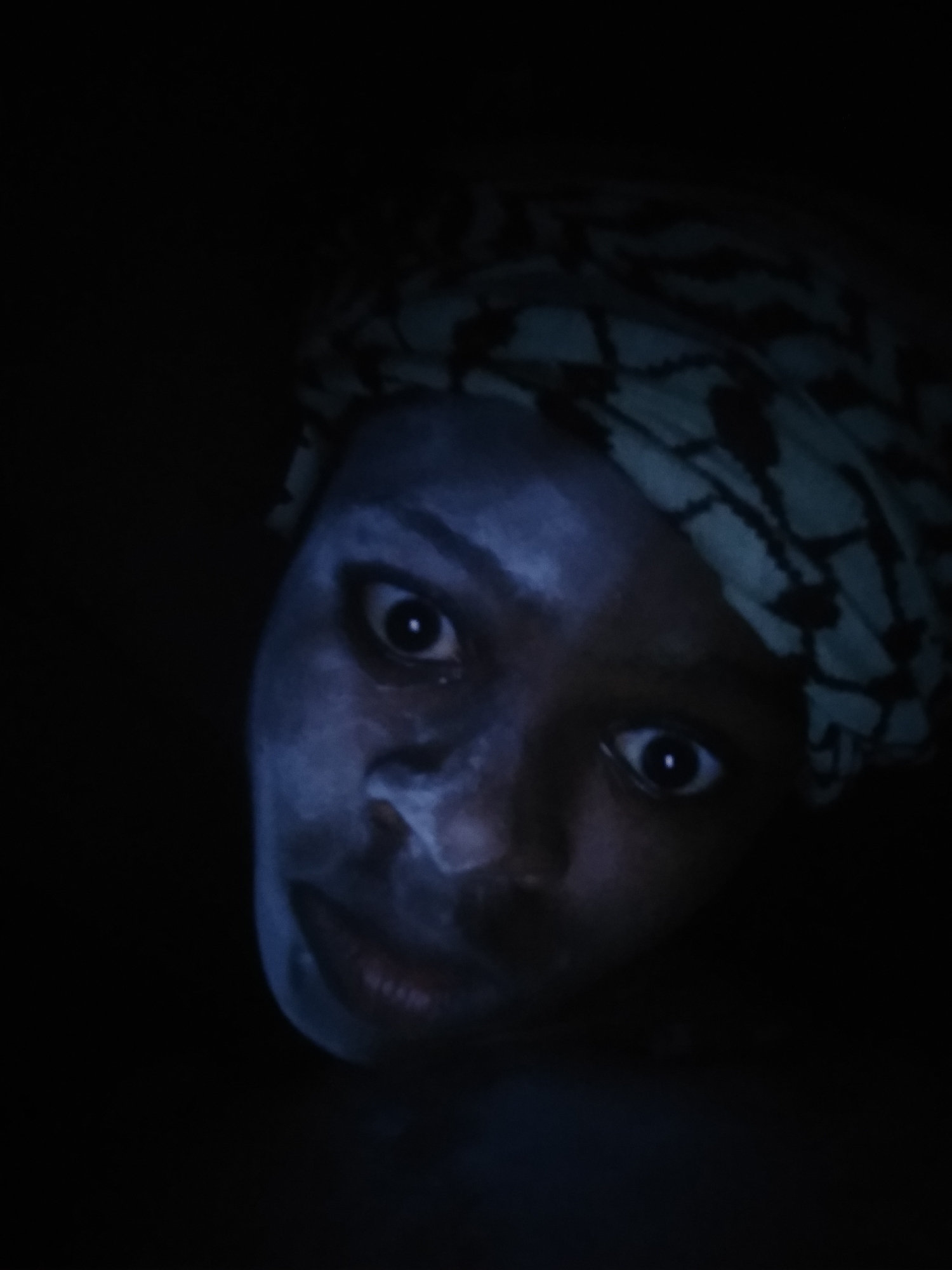 A person with light skin, possibly makeup or a mask, is wearing a patterned head covering. The background is dark, and the person is looking directly at the camera with a slightly tilted head. There is no indication of Shangisha, Nigeria, in the image.