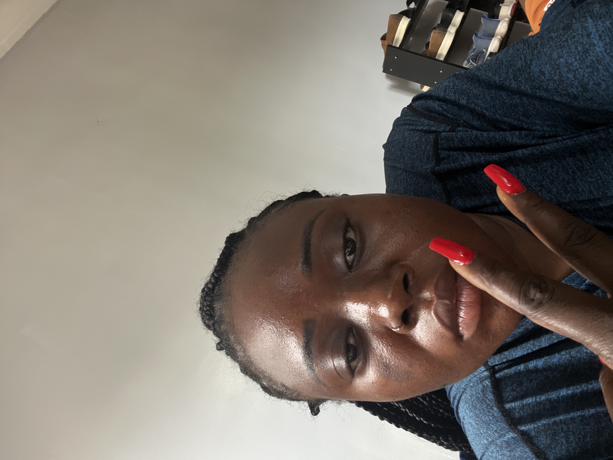 A close-up selfie of a dark-skinned person with black braids, facing the camera at an angle, with a blank white wall in the background. The person is wearing a blue textured shirt. The fingers of the person's right hand are raised, displaying bright red, rounded nail polish. A shoe rack with various shoes is visible blurred in the upper right corner of the image. The location or presence of Kubwa, Nigeria, cannot be verified from this image alone.