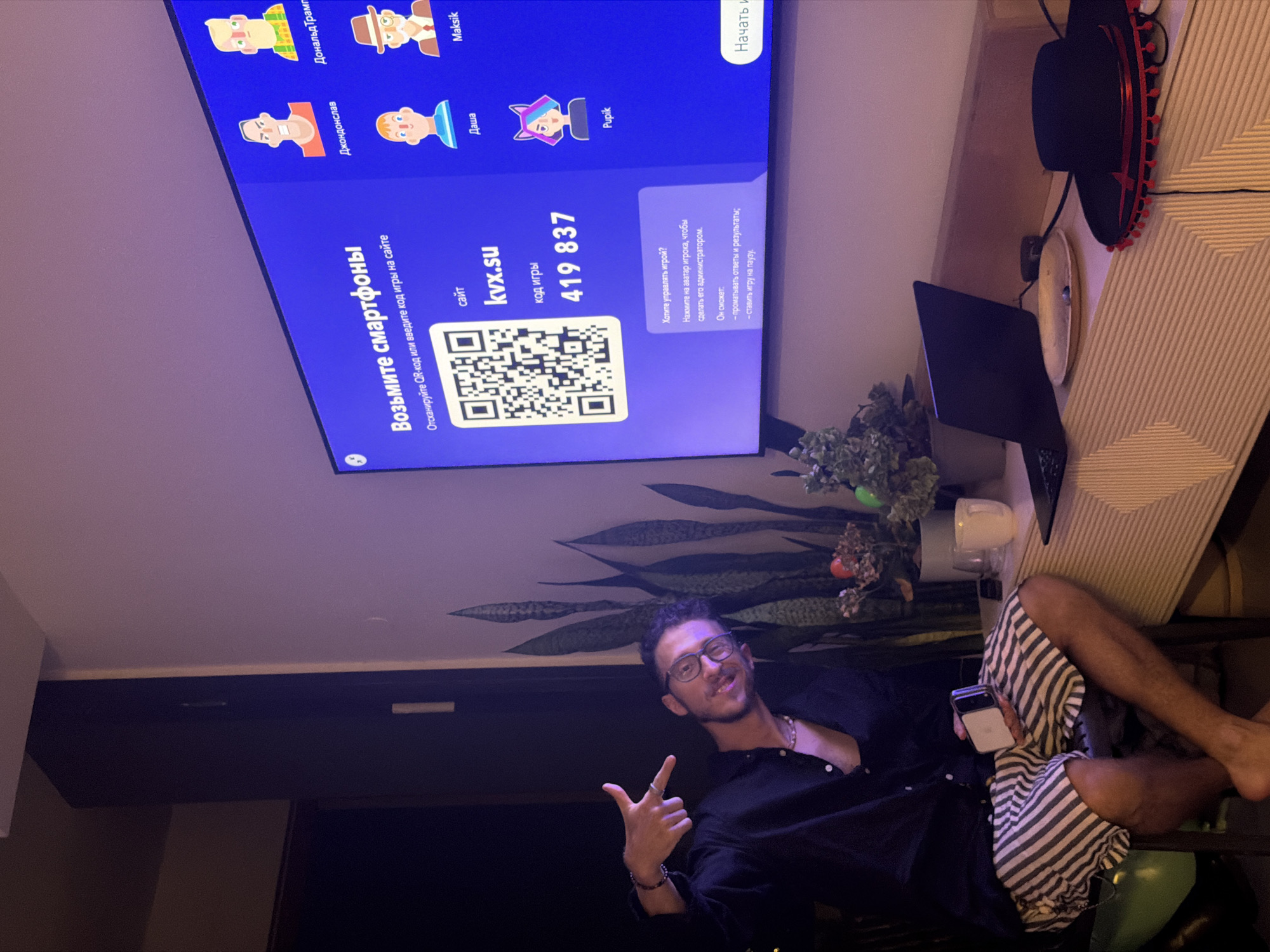 The image depicts an indoor scene, likely a living room or entertainment area. In the foreground, a person is seated on a chair, wearing striped shorts and a black button-down shirt, with their left hand pointing towards a large television screen. They are wearing glasses and a necklace. The television screen displays a QR code, a website address "kvx.su", and a game code "419 837". Various avatars with names are also visible on the screen, suggesting a gaming or interactive platform. On a surface below the television, there is a laptop, a hat with red trim, and some plants. The lighting suggests it is nighttime or a dimly lit environment. The presence of a phone in the person's hand and the overall setup indicates an engagement with the displayed content on the television. There are no specific visual cues that directly indicate the city of Tel-Aviv, Israel.