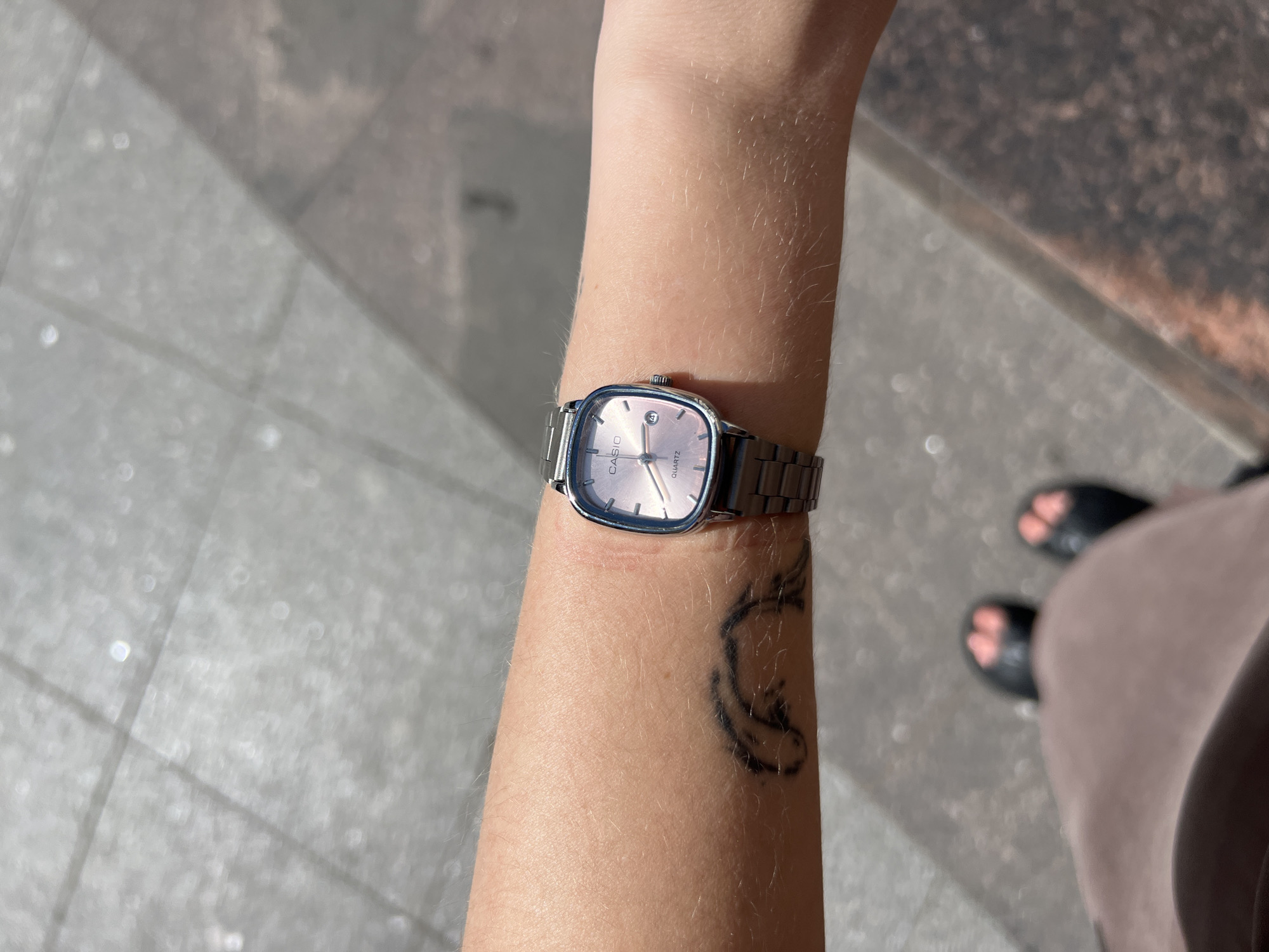 A close-up, high-angle shot shows a person's left arm with a silver wristwatch on their wrist. The watch has a square face and a metal band. Below the watch, there is a black tattoo on the arm. The arm is resting on a textured, grey surface that appears to be cobblestone or pavement, suggestive of an outdoor urban environment. In the lower part of the frame, the tops of two black sandals with orange accents are visible. The background is blurred but suggests an outdoor setting with sunlight casting shadows. The image appears to have been taken in Saint Petersburg, Russia, based on the typical urban pavement and outdoor lighting conditions often found in the city during sunny weather.