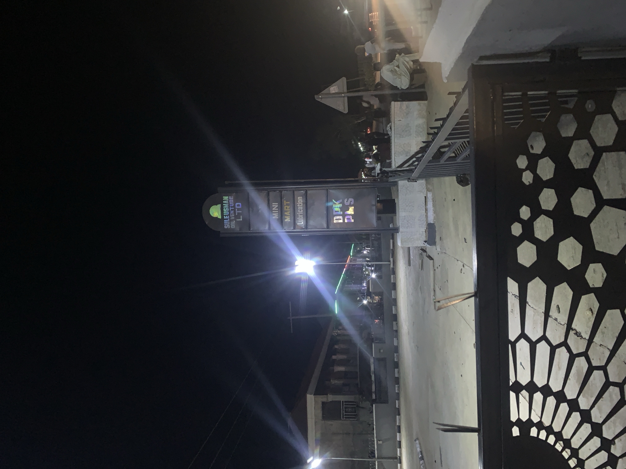 At night in Jalingo, Nigeria, a prominent sign displays "SULE USMAN OIL VENTURE LTD," with additional text indicating "MINI MART" and "Lubrication." Below this, "DPK PMS" is visible. The sign is illuminated and positioned in front of a building with a covered walkway supported by columns. Streetlights cast bright beams across the scene, and the sky is dark. Several individuals are present, some sitting near a railing and others standing. A dark metal gate with a honeycomb pattern is in the foreground. The area appears to be a commercial establishment, possibly a gas station or convenience store.