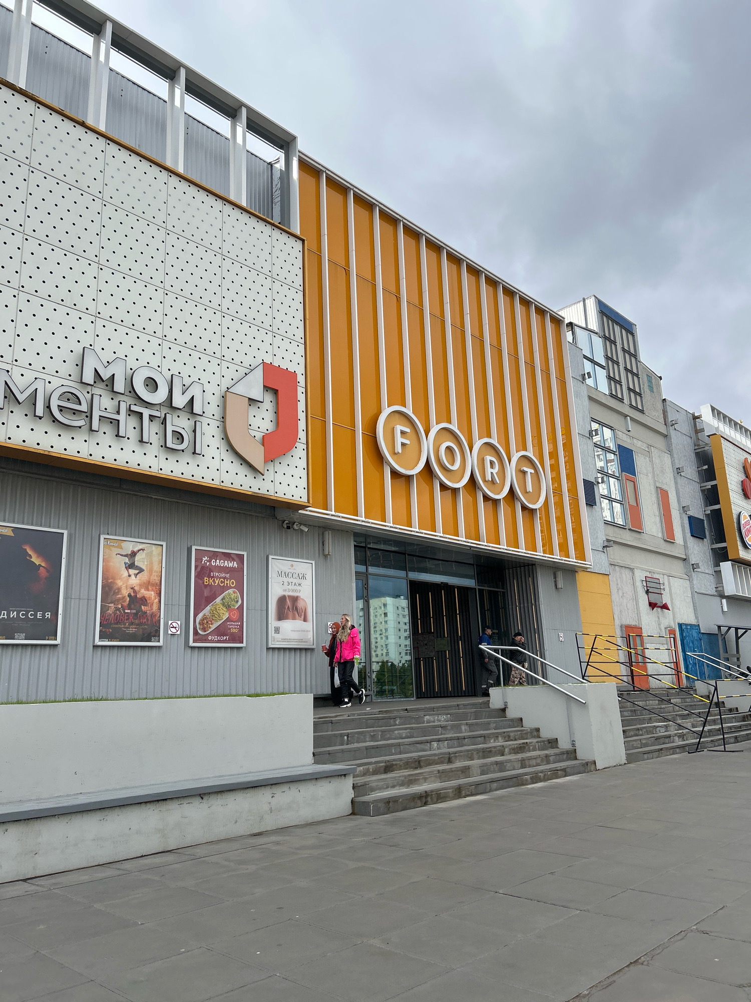 A low-angle, full-shot, exterior photograph captures the facade of the "FORT" cinema and "МОИ МОМЕНТЫ" shopping center on a cloudy day. The building features a prominent orange paneling with vertical white stripes, contrasting with a white facade adorned with a regular pattern of small black dots. Large, circular white lettering spells out "F O R T" above the entrance. Below the "МОИ МОМЕНТЫ" signage, four movie posters are displayed: "ДИССЕЯ", "ЧЕЛОВЕК-ПАУК", "GAGAWА ВТРОЙНЕ ВКУСНО", and "МАССАЖ". Two individuals are visible in front of the entrance, one wearing a bright pink jacket and black pants, the other in darker clothing. Several other people are partially visible near the entrance and on the stairs. A Burger King sign is visible further down the building to the right. The foreground shows a paved walkway and concrete stairs leading up to the building. The architectural style appears modern and commercial.