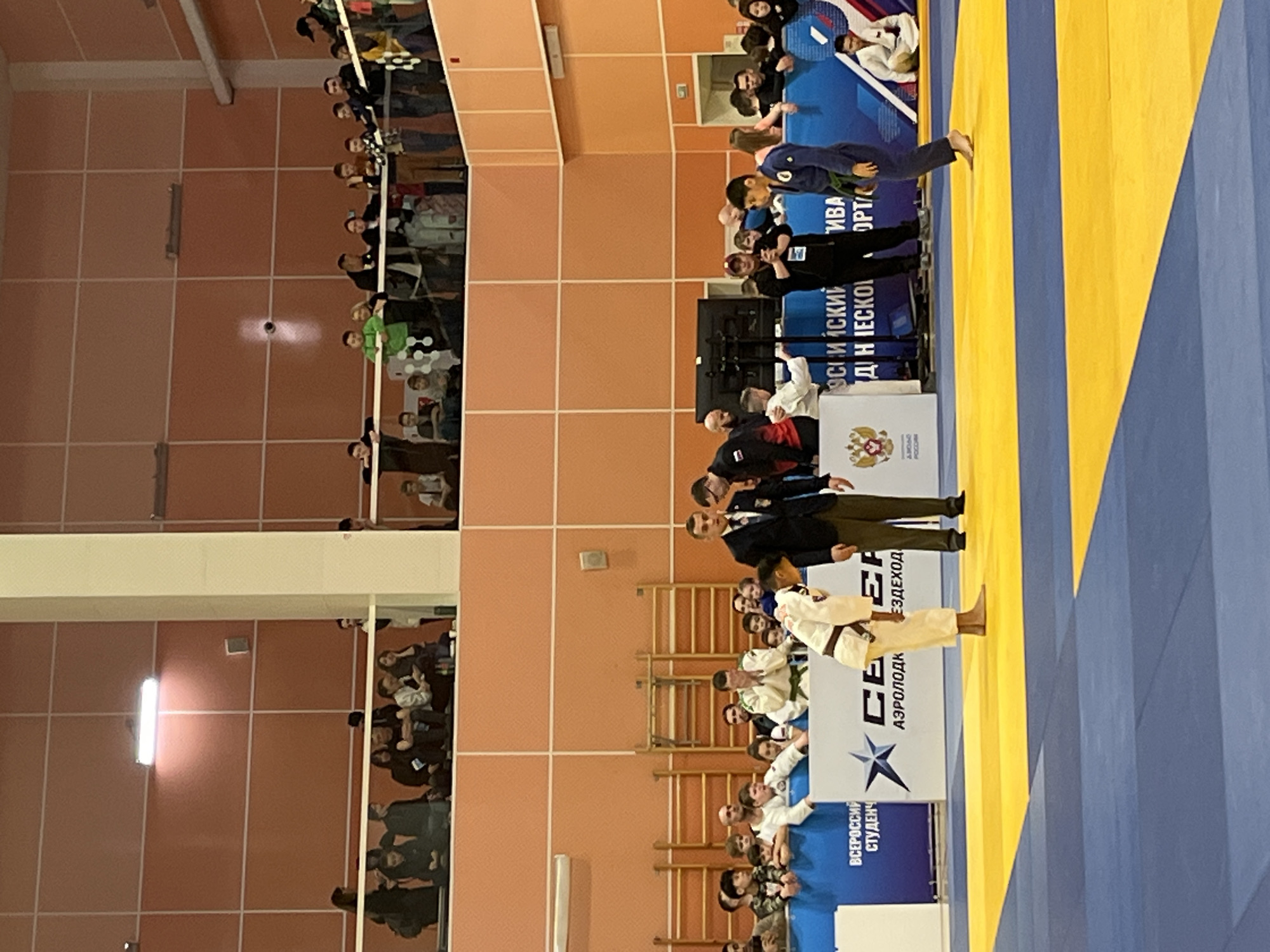 Here's a description of the image:
**Scene:** An indoor judo competition is taking place.
**Location:** The setting is likely in Krasnoyarsk, Russia, indicated by the “РОССИЯ” text and the Russian coat of arms seen on the banner.
**People:** Two judokas (one in a white uniform and the other in blue) are standing on the mat. Referees in black suits and officials are also present. Spectators can be seen in the stands.
**Subjects:** The focus is on the judo competition with the athletes and officials.
**Objects:** A judo mat (blue and yellow), banners with Russian text and the Russian coat of arms, folding tables, and chairs.
**Actions/Interactions:** The judokas appear to be bowing, likely at the start or end of a match. The referees are standing by to supervise.
**Environment:** The indoor arena has a tiled wall in the background. It is well-lit.
