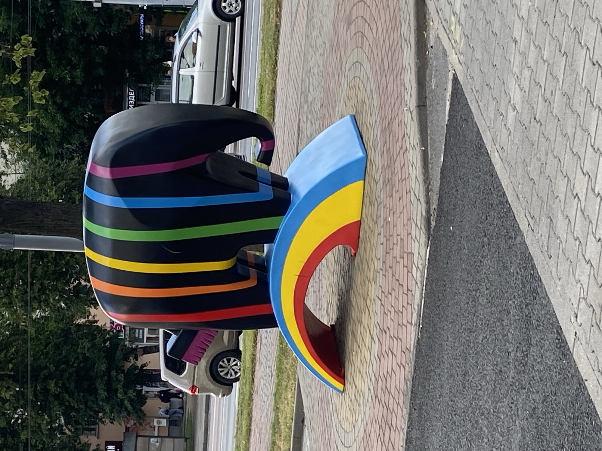 This is a vertical, eye-level, medium shot of a colorful sculpture on a city sidewalk. The sculpture is black with horizontal stripes in various rainbow colors, including pink, blue, green, yellow, orange, and red. It appears to be abstract or stylized, possibly representing an animal like an elephant or a mythical creature. Part of the sculpture curves upwards, forming a blue, yellow, and red arch.
The sculpture is situated on a paved sidewalk made of light-colored interlocking bricks laid in a circular pattern. To the left of the sculpture is a patch of dark asphalt, suggesting it's near a road or parking area. Behind the sculpture, there is greenery from trees and bushes, indicating an outdoor urban setting. Further in the background, a white car is parked on what looks like a street, and a few buildings are visible. The lighting suggests it is daytime. There is a small sign with Cyrillic text visible on the building in the background, but it is illegible. The scene appears calm and ordinary, with no specific activity or event taking place beyond the presence of the sculpture. The location context provided is Brest, Belarus.
