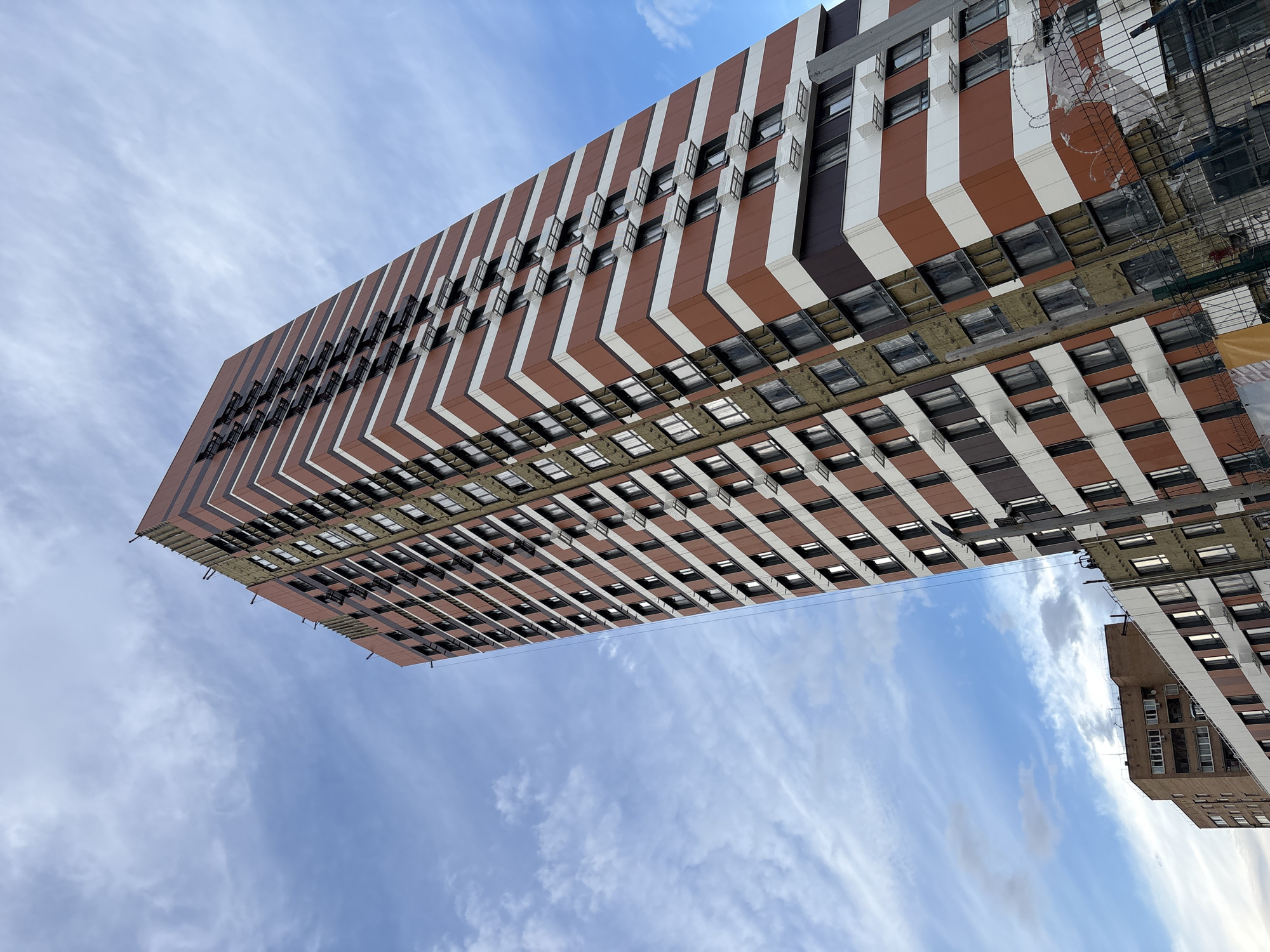 This is a low-angle, wide shot of a modern, tall residential building under construction. The building features a striking facade with alternating vertical bands of reddish-brown and white. Many windows are visible, some with open vents. The underside of the building reveals unfinished construction elements, including insulation and exposed framing. To the right, a portion of another, older brick building is visible, also with windows. The scene is set against a bright blue sky with wispy white clouds, suggesting it is daytime. The lighting indicates a sunny day. The overall impression is one of urban development and ongoing construction. No people are visible in the frame. No text is legible. The location context is Tarasovka, Russia.