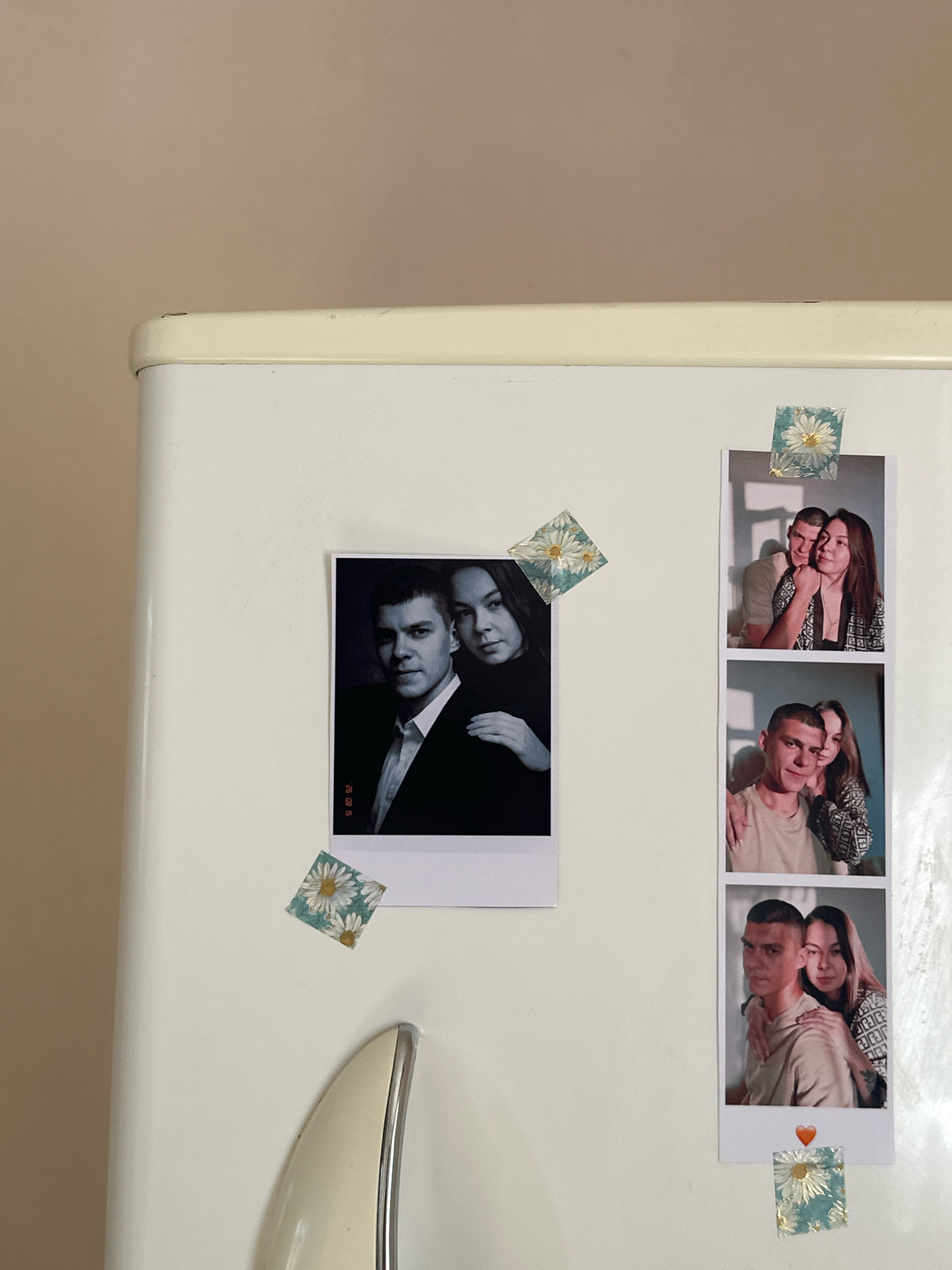 The image shows a close-up of a white refrigerator door. Several photographs are attached to the door with magnetic decorative elements. On the left side of the refrigerator door, a large, rectangular photograph features a man in a suit and a woman. To the right of this photograph, a strip of three smaller, vertical photographs are displayed. The top and bottom photographs in this strip show couples embracing. The middle photograph in this strip also depicts a couple. Each of these smaller photographs is topped with a square decorative element featuring a floral pattern. The bottom photograph in the strip has a small heart-shaped magnet beneath it. Another square decorative element with a similar floral pattern is attached to the refrigerator door between the large photograph and the strip of smaller photographs. The refrigerator door is white and appears to be clean. The background of the image is a plain, light brown wall. The scene does not provide any specific visual cues or landmarks to indicate its location in the city of Gyumri, Armenia.