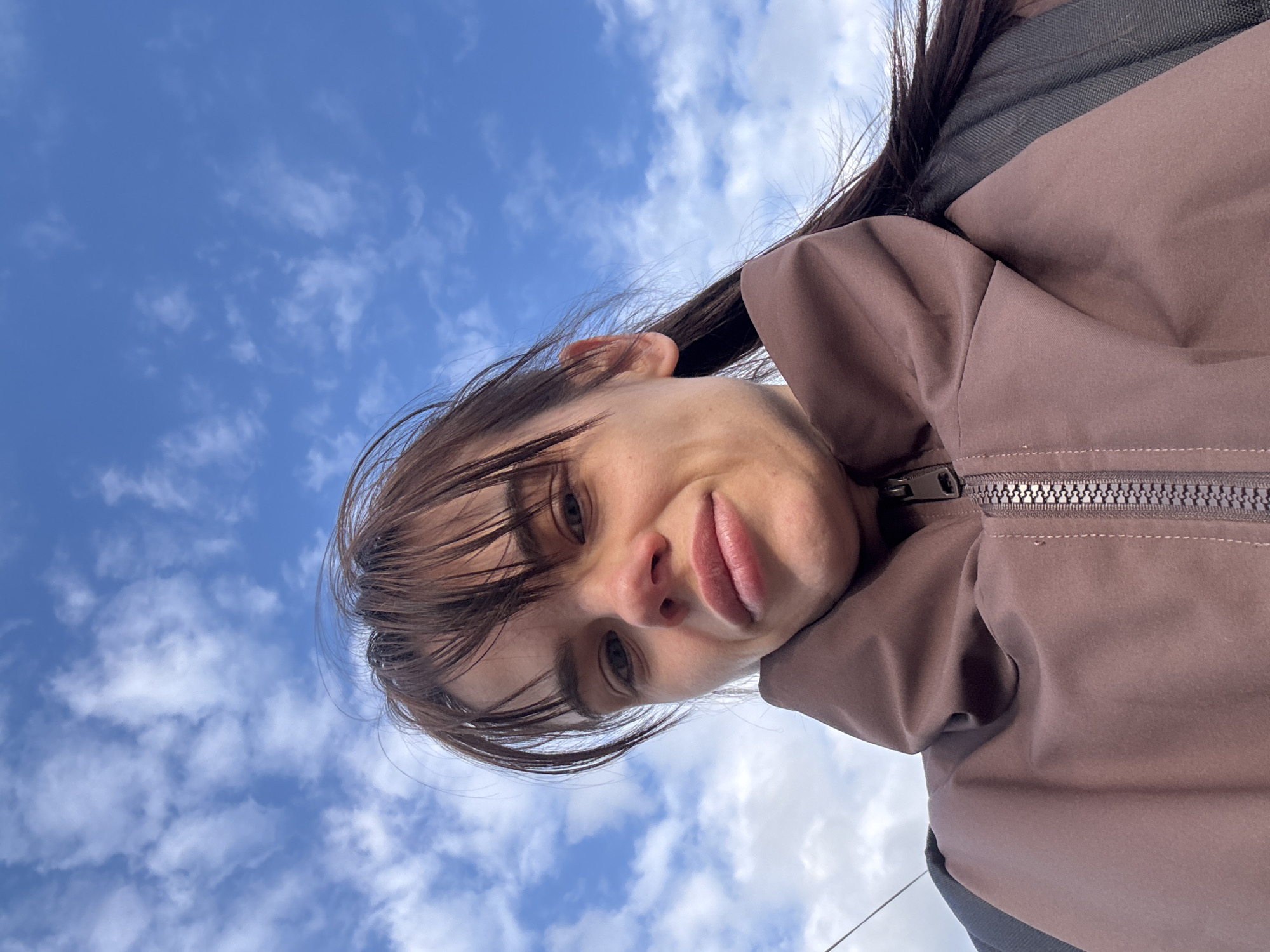 An outdoor, low-angle portrait captures a person, likely female, looking directly into the camera. The subject's face is prominent, featuring dark hair with bangs, and a slightly pouting or quizzical expression. She is wearing a muted brown or reddish-brown jacket with a prominent silver-toned zipper. A dark grey or black strap, possibly from a backpack or a jacket element, is visible over her right shoulder.
The background consists of a vibrant blue sky dotted with scattered white, cumulus clouds, indicating bright daylight. The overall lighting is natural and even. The image is taken from a perspective looking upwards at the subject, emphasizing the expansive sky above. There is no visible text in the image. The scene is set in an open-air environment in Rostov-on-Don, Russia.