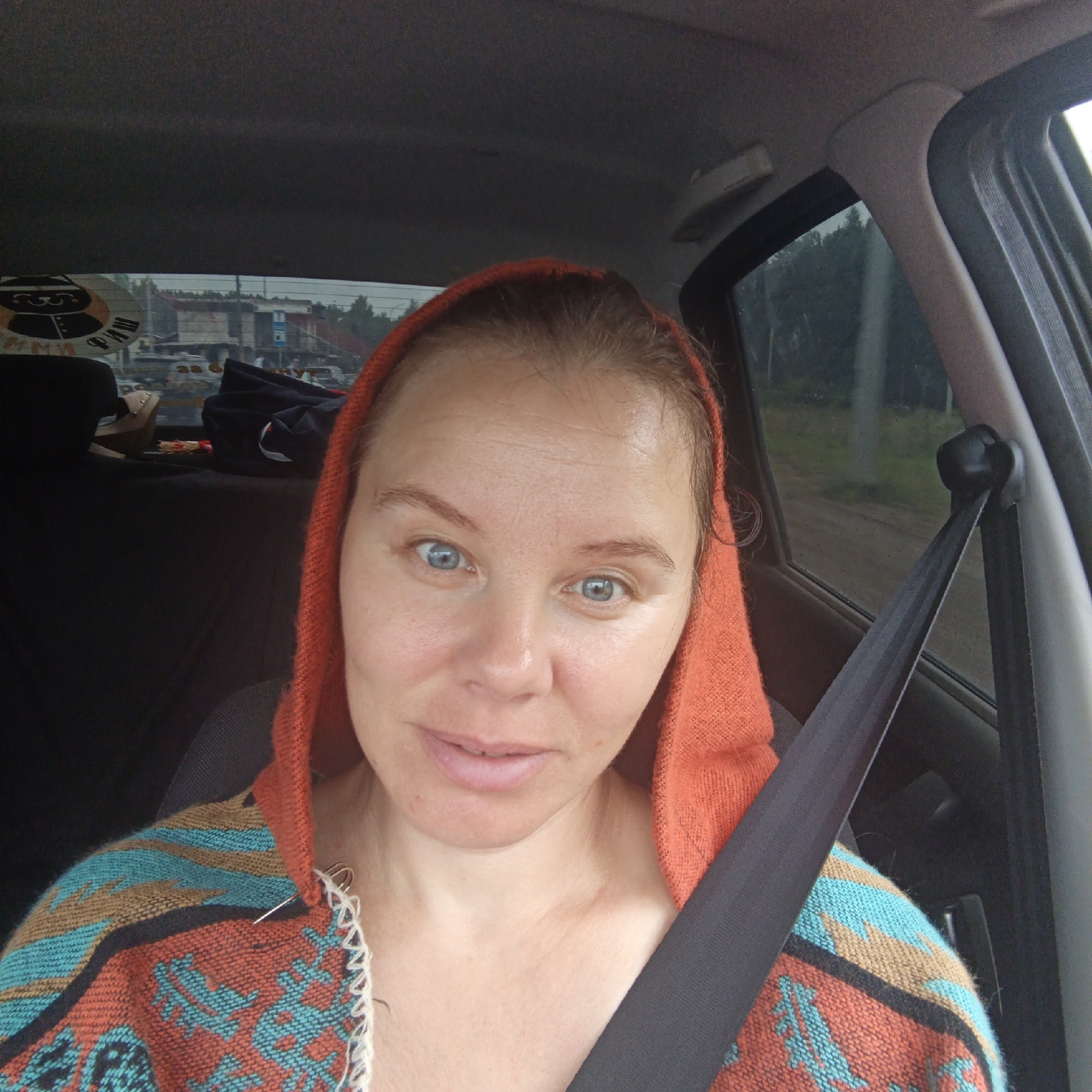 The image shows a woman in the driver's seat of a car, wearing an orange hooded garment with a patterned poncho over it. She is looking at the camera, and a seatbelt is visible across her chest. Through the car window, a rural or suburban landscape can be seen, with trees and some buildings in the distance. The style of the buildings suggests a location in Russia. There are no specific landmarks visible that would definitively place this image in Chaikovsky.