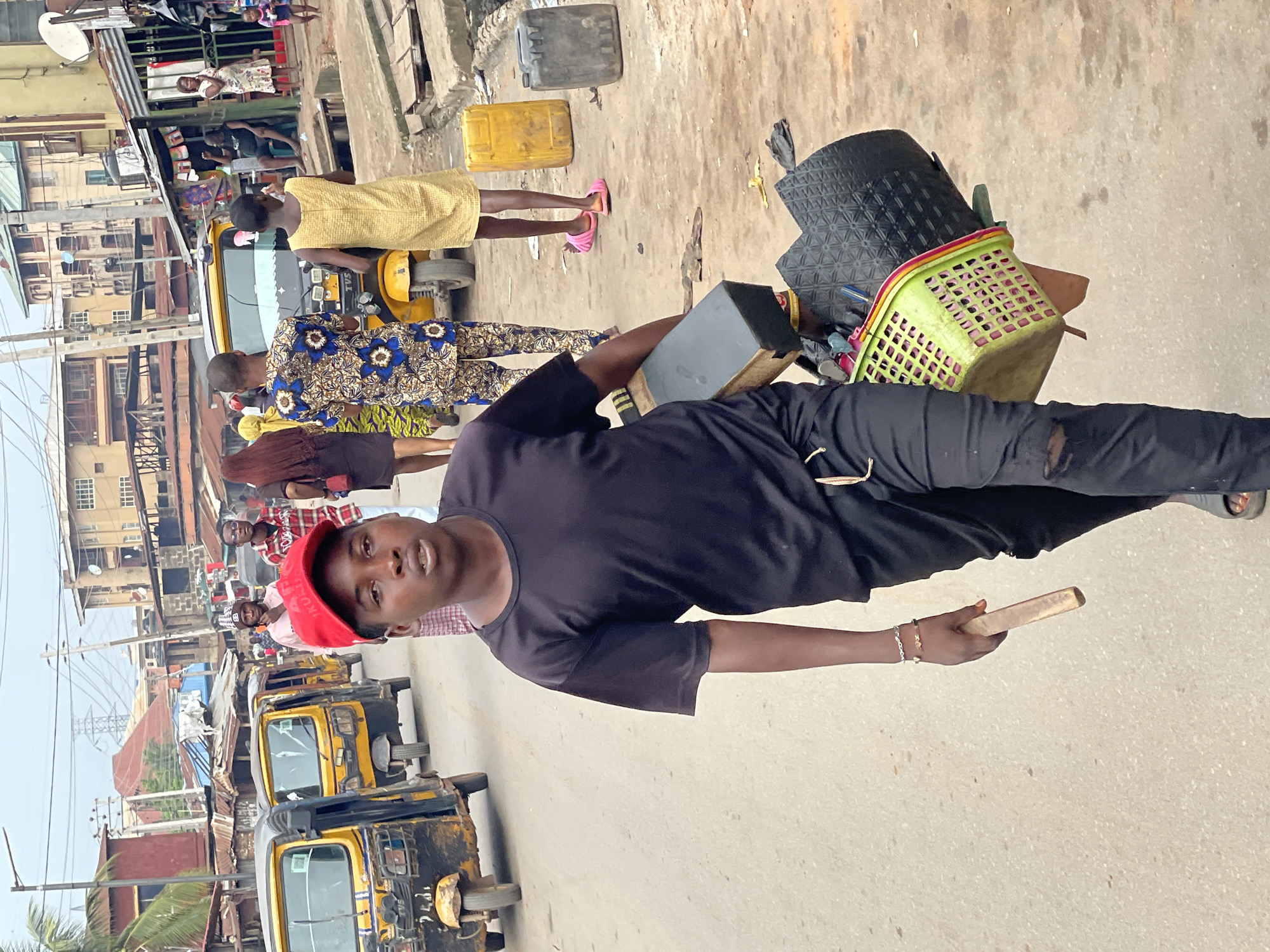 The image shows a man in a black t-shirt and black pants, wearing a red cap, standing on a concrete road in Onitsha, Nigeria. He holds a wooden object. A green basket and a black mat are on his side. In the background, several yellow auto rickshaws (keke) are parked on the side of the road, and pedestrians walk along the side of the street. Several buildings are visible along the side of the road.