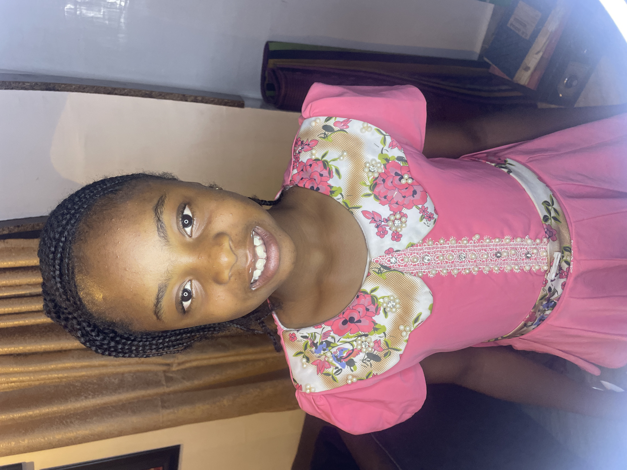 A young girl is shown in a brightly lit, indoor environment. She is smiling and facing the camera. The girl has dark skin and her hair is braided. She is wearing a pink dress with a white floral pattern around the neckline, which is decorated with small pearl-like embellishments. The background consists of a wall and some furniture covered in brown and dark fabrics, and a shelf or table area with what appears to be books and other items. There is no visible evidence to confirm this is Jos, Nigeria.