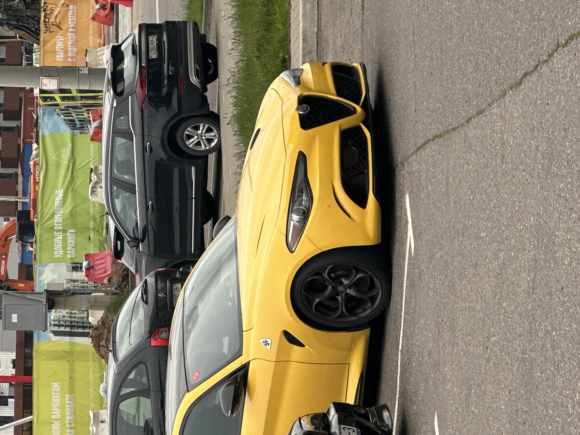 Here is a forensic description of the image:
The image shows a yellow Alfa Romeo Giulia parked on a street in Бараўлянскі сельскі Савет, Belarus. A black SUV is parked behind it. In the background there are construction signs and what looks like a construction site. The light conditions suggest it is daytime.