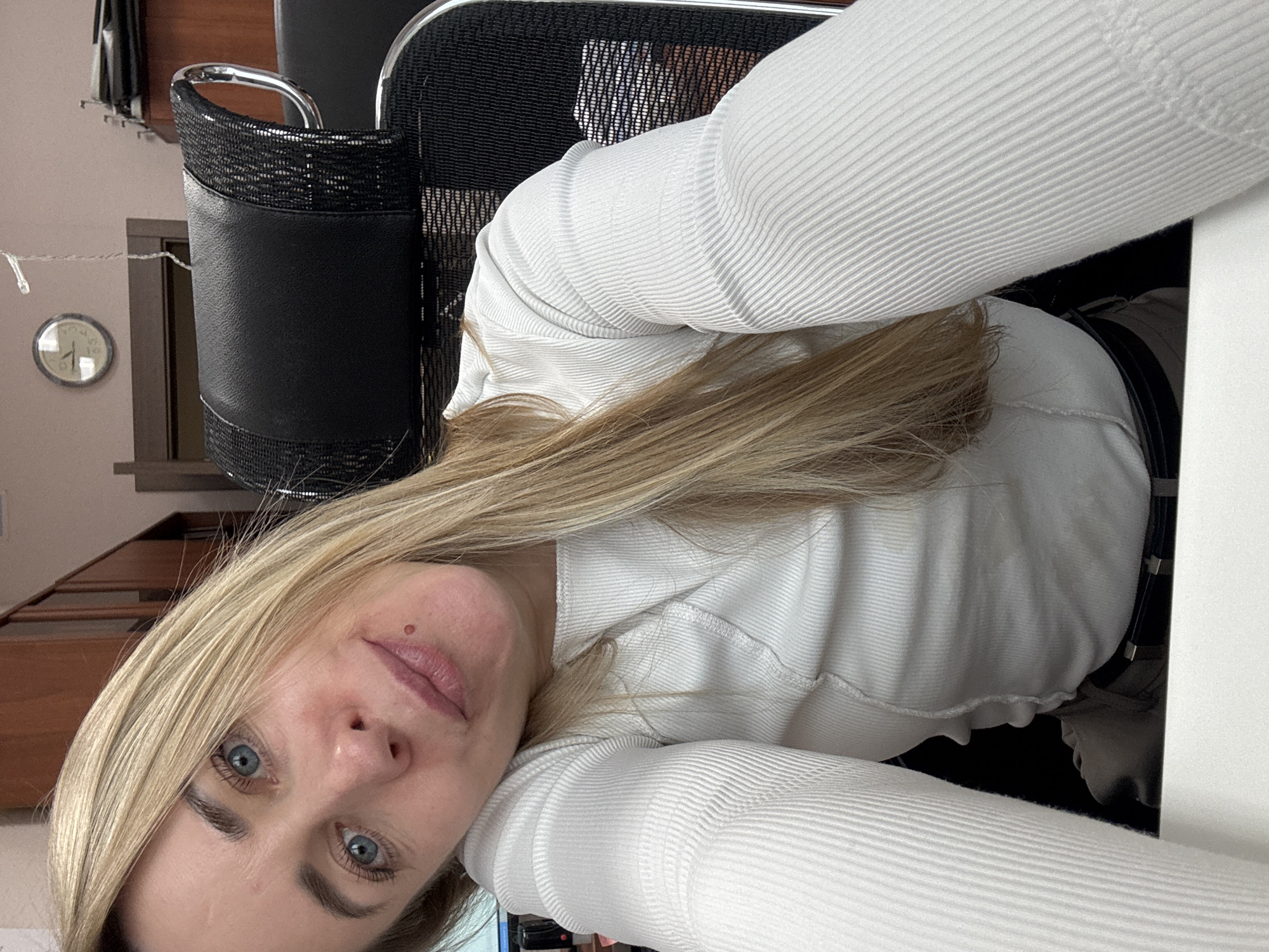 A selfie of a woman with blonde hair and blue eyes. She is wearing a white ribbed long-sleeved shirt. The background includes part of an office chair and a glimpse of a room with a clock on the wall and wooden shelving. The location is Chaikovsky, Russia.