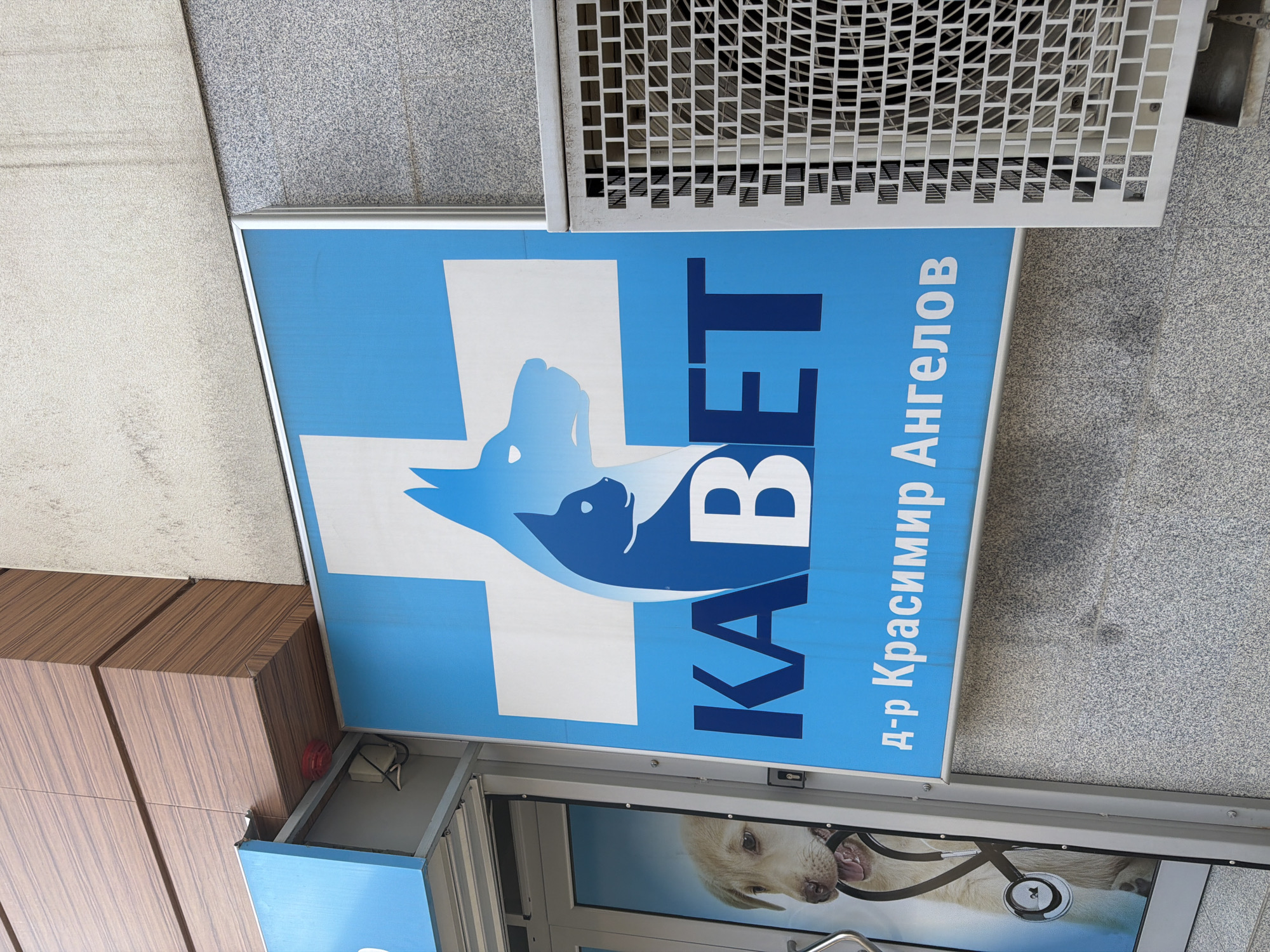 The image shows a sign for a veterinary clinic, presumably in Plovdiv, Bulgaria. The sign has a light blue background with a white medical cross. In the center of the cross is a stylized image of a dog and a cat, both in a darker blue. To the right of the cross, vertically stacked, are the words "КАВЕТ" in large, dark blue letters, followed by "д-р Красимир Ангелов," also in dark blue. The sign is mounted above a glass door/window, below which can be seen an image of a dog wearing a stethoscope. The building facade is a mix of materials including wood-like panels, granite, and metal grating.