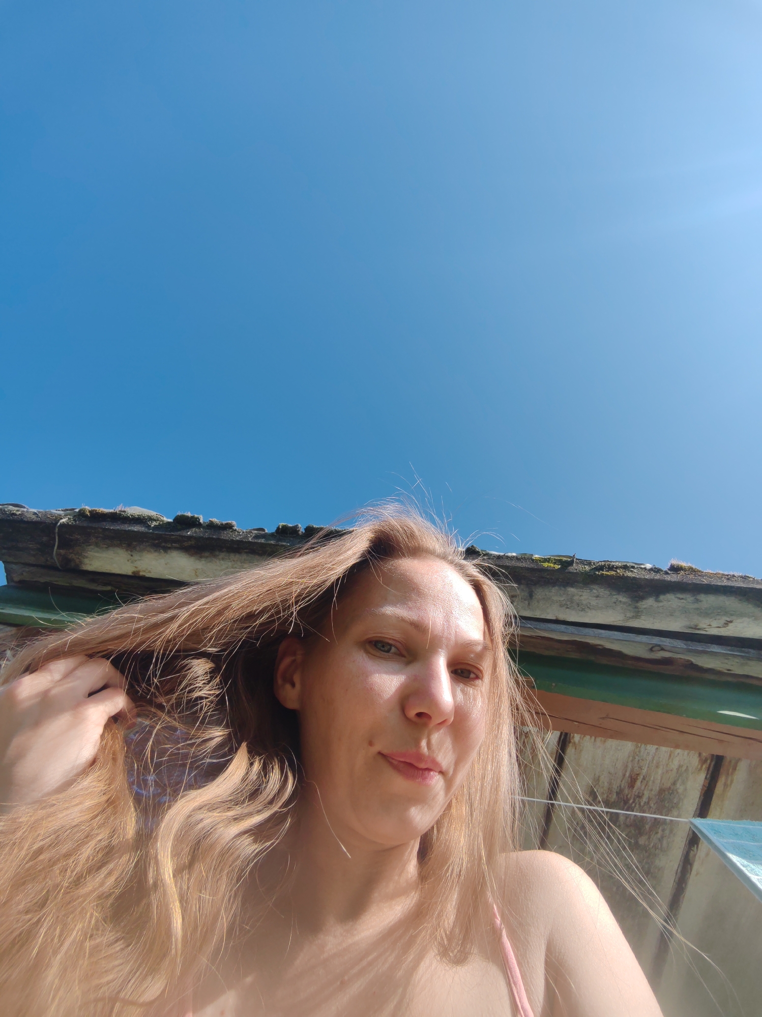 A low-angle, close-up outdoor selfie shows a woman with blonde hair holding a section of her hair with her left hand. She is wearing a pink top. Her head is tilted slightly to her right, and her gaze is directed towards the camera. Sunlight illuminates her face, casting shadows and highlighting freckles. Behind her, the roof of a wooden structure is visible, with weathered planks and some moss growing on it. The sky above is a clear, bright blue. The scene appears to be set on a sunny day in Чухлэм, Russia.
