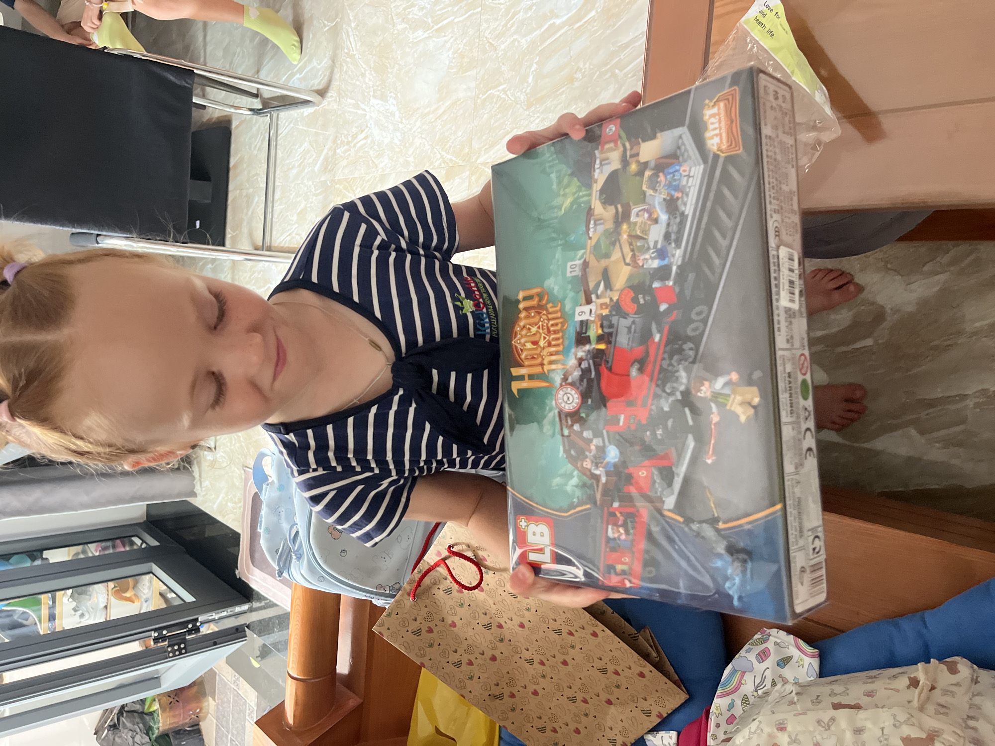 A young girl with braided blonde hair is holding a box of "Harry Magic" branded building blocks, likely a toy set. She's wearing a blue and white striped shirt. The scene takes place inside a room with a tiled floor, wooden furniture, and a window. Visible in the background are a chair with a black seat and the feet of another person wearing yellow socks. Also in the frame are a gift bag with heart patterns, a blue pillow, and a white patterned fabric. Based on the surroundings, the location may be a residential area in Bac Nha Trang Ward, Vietnam.