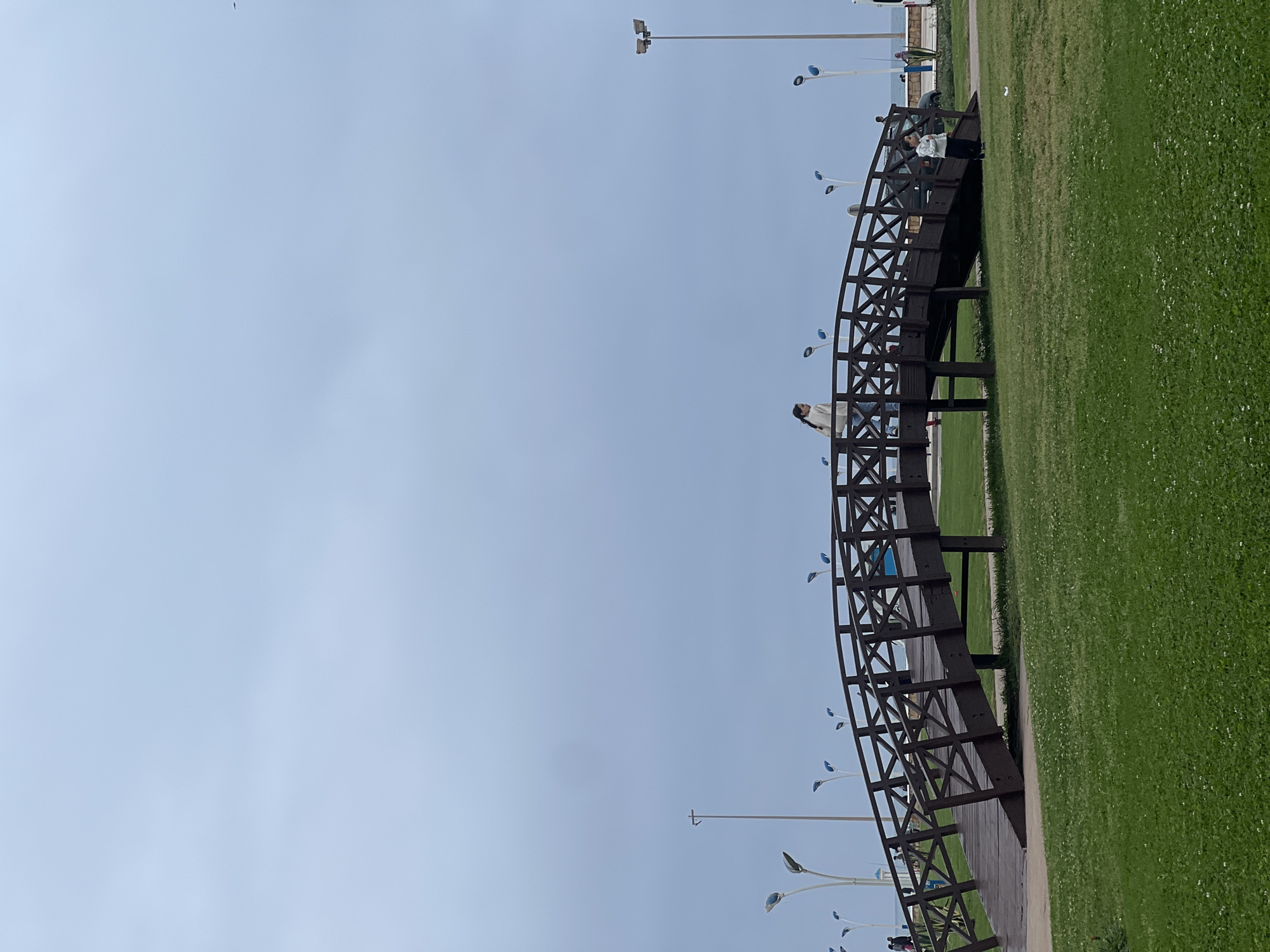 The scene depicts a curved, wooden footbridge located in M'Diq, Morocco. The bridge is situated on a strip of green grass, with the sky visible in the background. The bridge has a lattice-like design on its sides. On the bridge are several people. Streetlights are visible near the bridge. The ground near the bridge appears to be paved with wooden planks.