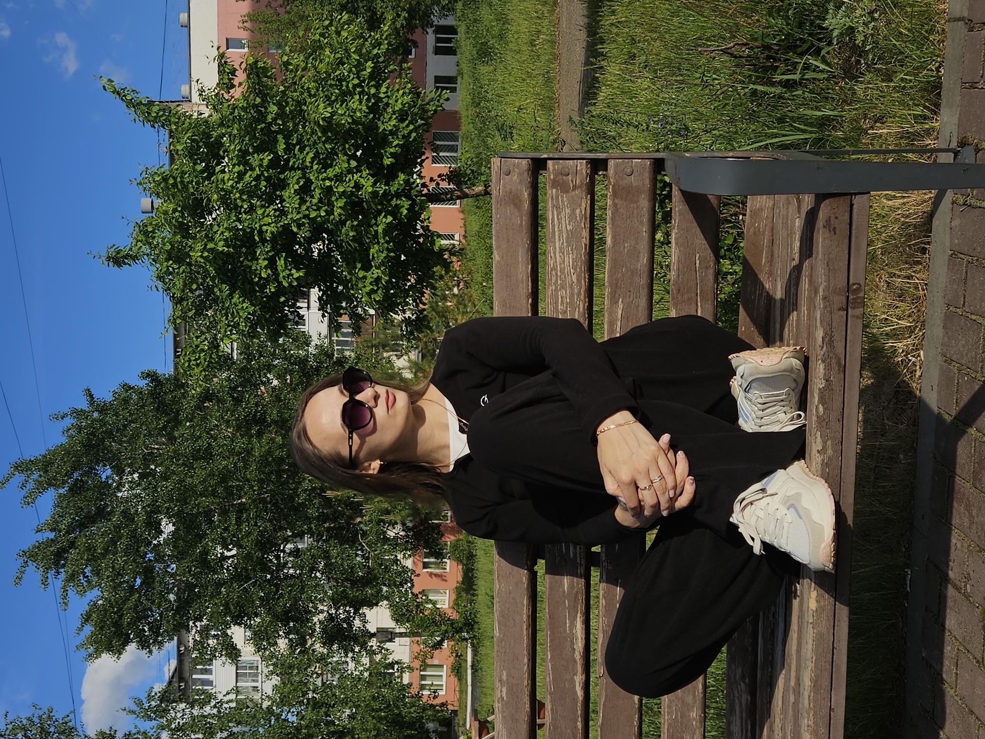 A young woman wearing sunglasses and black clothing is sitting cross-legged on a wooden bench outdoors. She has jewelry on her hands and wrists. The bench is in a park-like area with trees, grass, and a paved pathway. Buildings are visible in the background. The location is Chaikovsky, Russia.