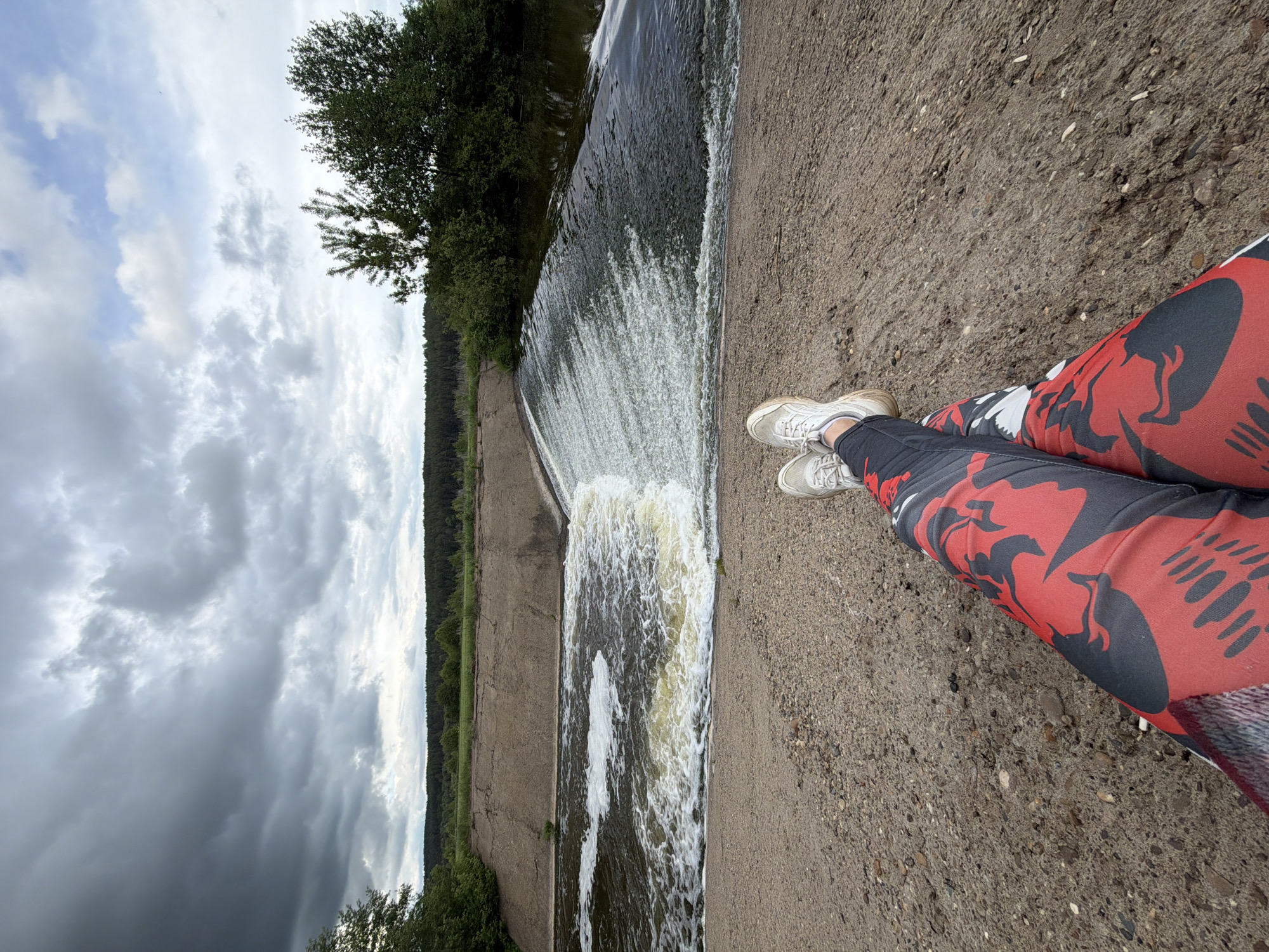 The photograph presents a scene at the location of the dam in Nizhnii Vishur, Russia. The view is from a high angle, looking down at the dam and the water flowing over it. An individual is present, with their legs extending into the frame, clad in red and black patterned pants and wearing white sneakers. The dam is constructed of concrete, and water is rushing over the edge. Beyond the dam, a line of trees and dense vegetation is visible along the riverbank. The sky is overcast with dark clouds.