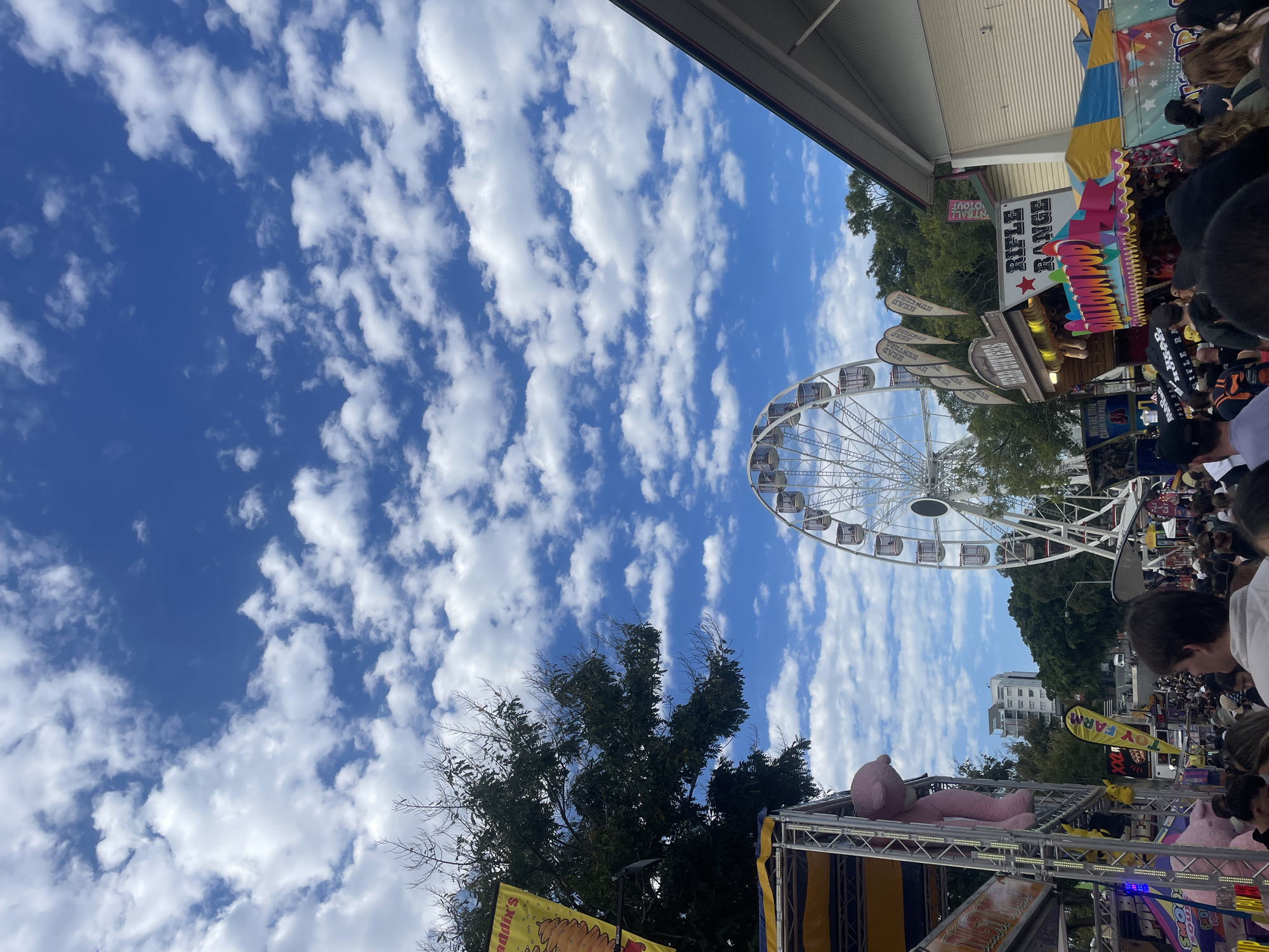 This image depicts a lively outdoor event, likely a fair or carnival, set against a backdrop of a bright blue sky dotted with fluffy white clouds. The scene captures a crowd of people milling around various attractions.
Prominently featured is a large white Ferris wheel, its structure rising towards the sky. To the left, a stall with a yellow awning displays a large, pink teddy bear hanging from its side. Adjacent to this, another stall has a banner that reads "SMOOTHIES." Further to the right, there are other stalls, one with a sign for "RIFLE RANGE" and another advertising "BALLOON POP." Flags with the text "HEAD HUNTERS" are visible, suggesting carnival games or attractions. The presence of trees and some buildings in the background indicates an urban or semi-urban setting.
A specific detail pointing to the location is a banner on a stall that reads "QUEENSLAND." Given the context of a fair with a Ferris wheel and carnival games, and knowing that Bowen Hills in Australia is a known location for such events, it is plausible this image was taken at the Bowen Hills Brisbane Ekka. However, without explicit signage confirming this, it remains an inference. The overall atmosphere is one of a bustling public gathering, filled with diverse individuals enjoying the activities.