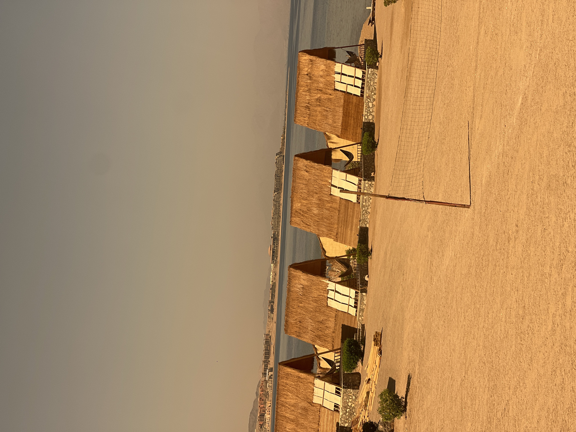 A bright, hazy daytime, wide-angle, eye-level shot captures a beachfront scene in Nuweiba, Egypt. In the foreground, a sand volleyball court with a net is visible. Beyond the court, a row of four small, thatched-roof bungalows with wooden frames are arranged along the shore. The bungalows are identical in design, each featuring two foldable, white canvas awnings at the front. Small green bushes are planted in front of and between the bungalows. A stack of wooden planks is to the left of the first bungalow, suggesting ongoing construction or maintenance. The sea stretches out behind the bungalows, meeting a distant, hazy shoreline with buildings that appear to be resorts or hotels. Mountains are visible in the far distance, shrouded in atmospheric haze. The sky is a pale, washed-out blue, indicating either a very clear but dusty atmosphere or a slight overexposure. There are no people visible in the frame.
